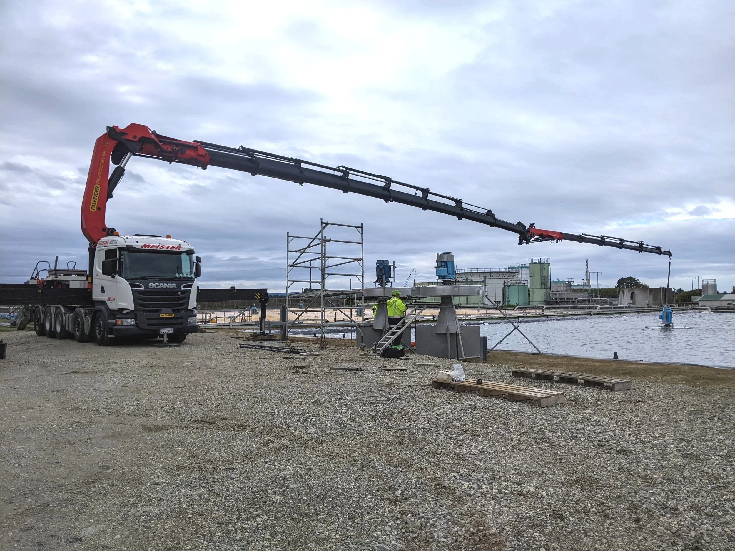 Construction site near a body of water with a large truck equipped with an extended crane arm, scaffolding, and workers wearing safety gear.