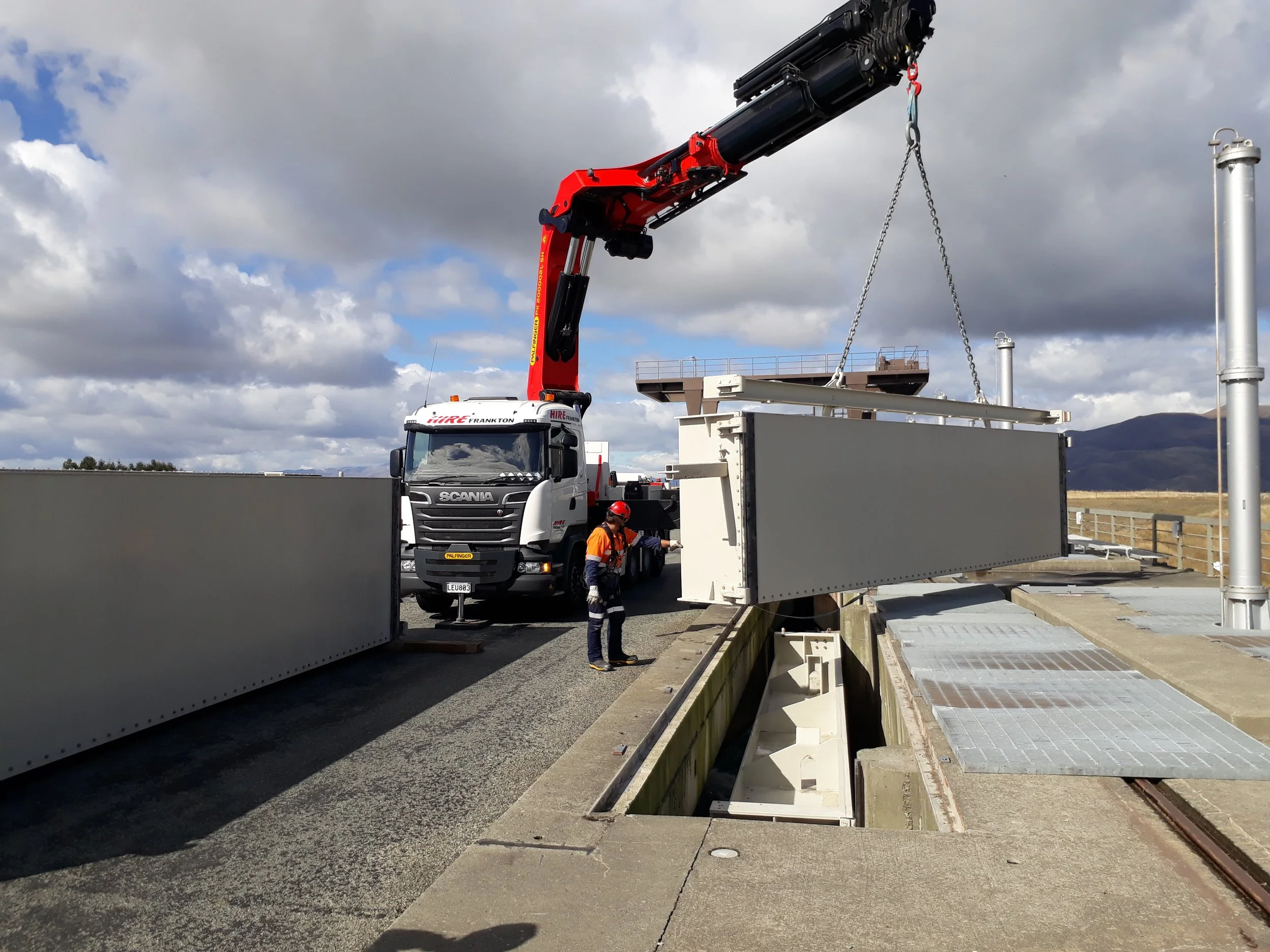A crane truck lifts a large, rectangular metal structure on a railway platform with a worker nearby, under a partly cloudy sky.