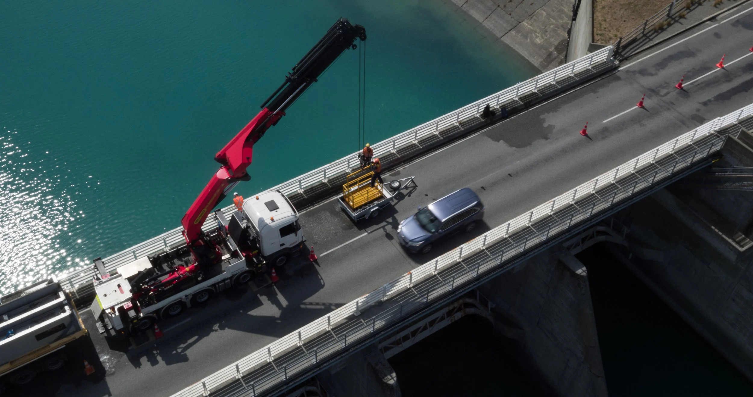 Construction workers on a bridge installing or repairing safety barriers using a crane truck. There are orange traffic cones guiding traffic, and a gray vehicle passing by. The bridge spans over a body of water.