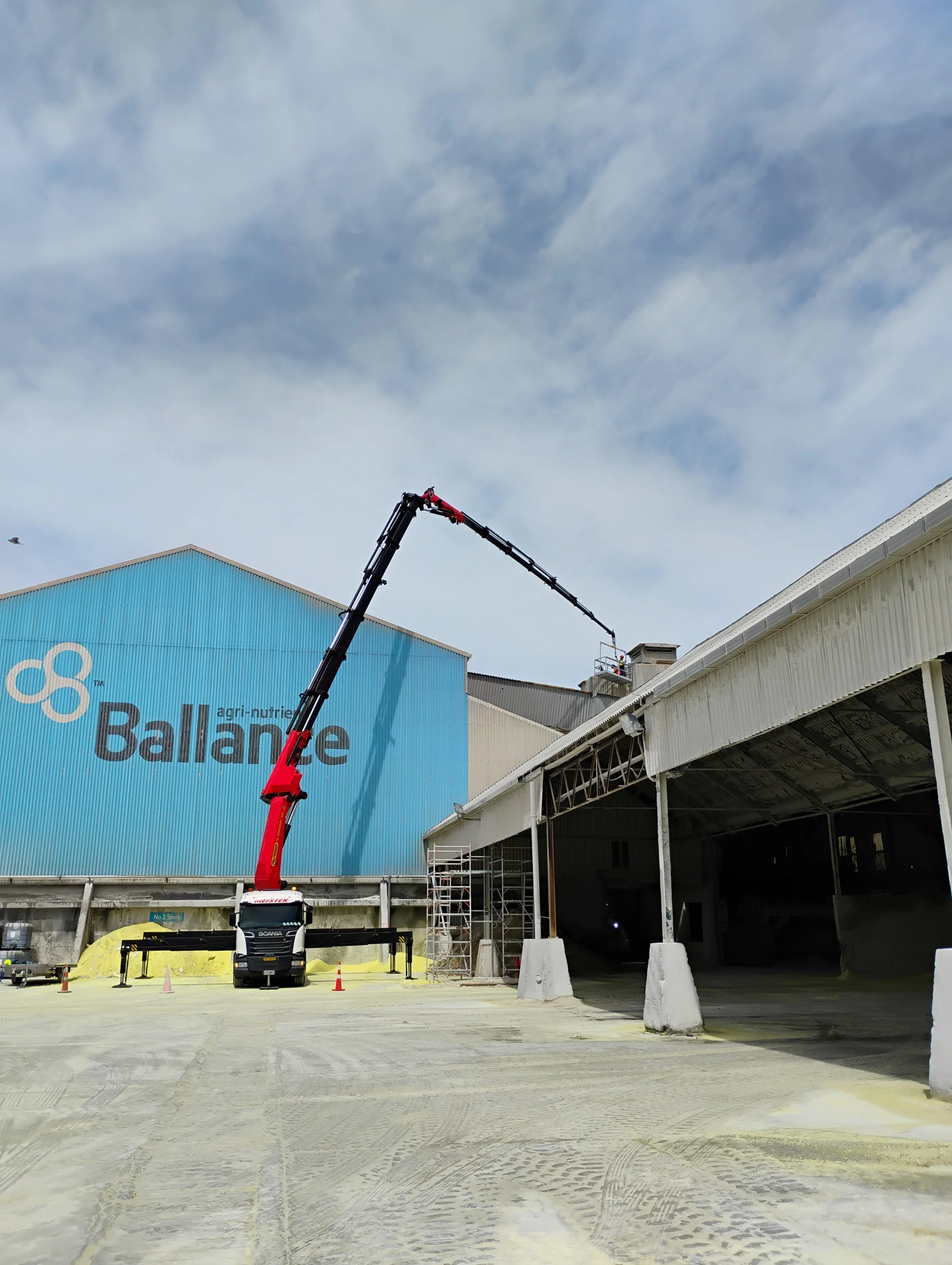 Construction site with a large truck crane extending a long arm, near a building with a blue wall that has the words 'agri-nutrition Balance'. The sky is partly cloudy, and the area has construction cones and scaffolding.