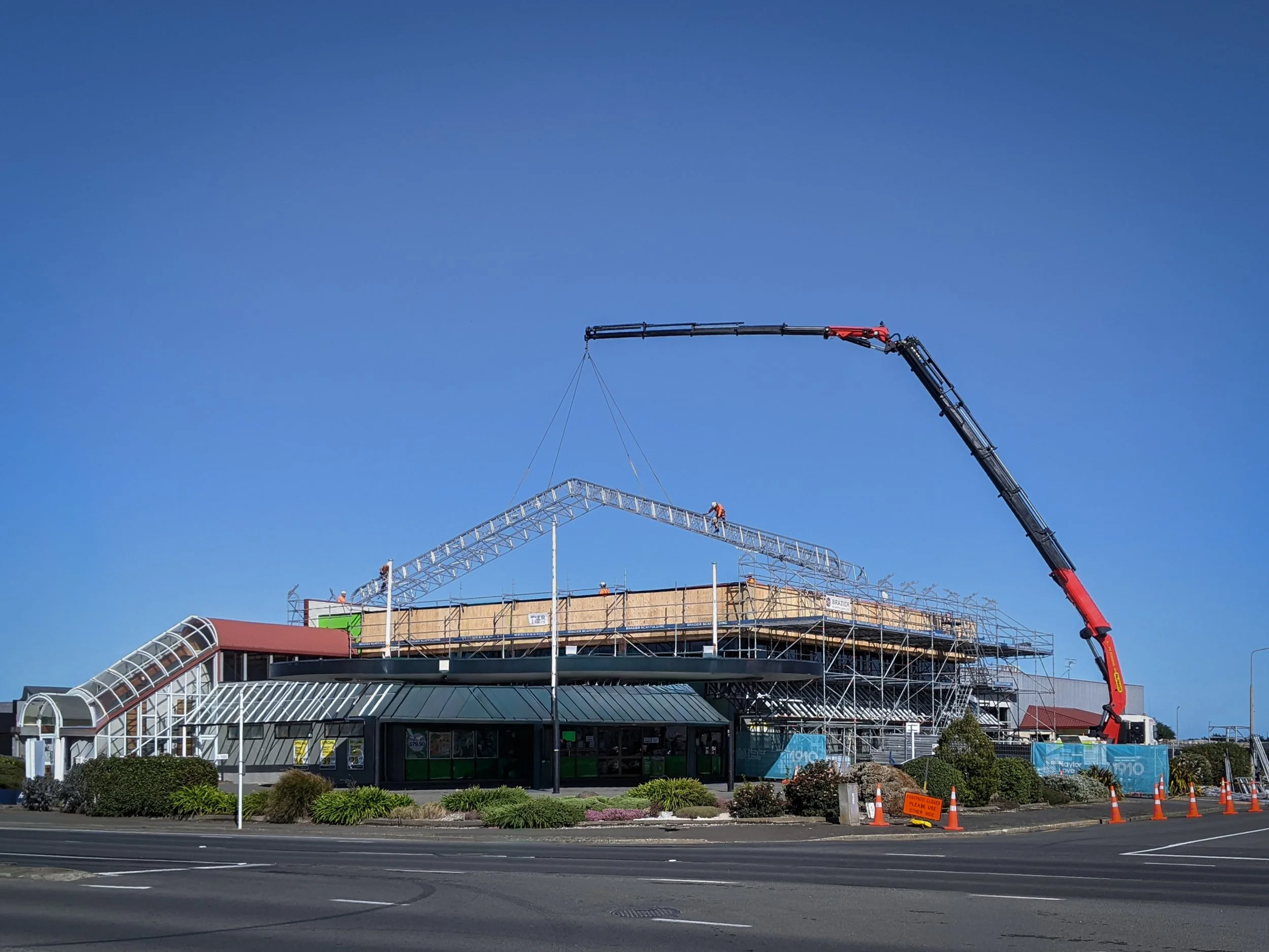 Construction site with a building under construction, scaffolding, a crane, and workers installing a metal structure against a clear blue sky.