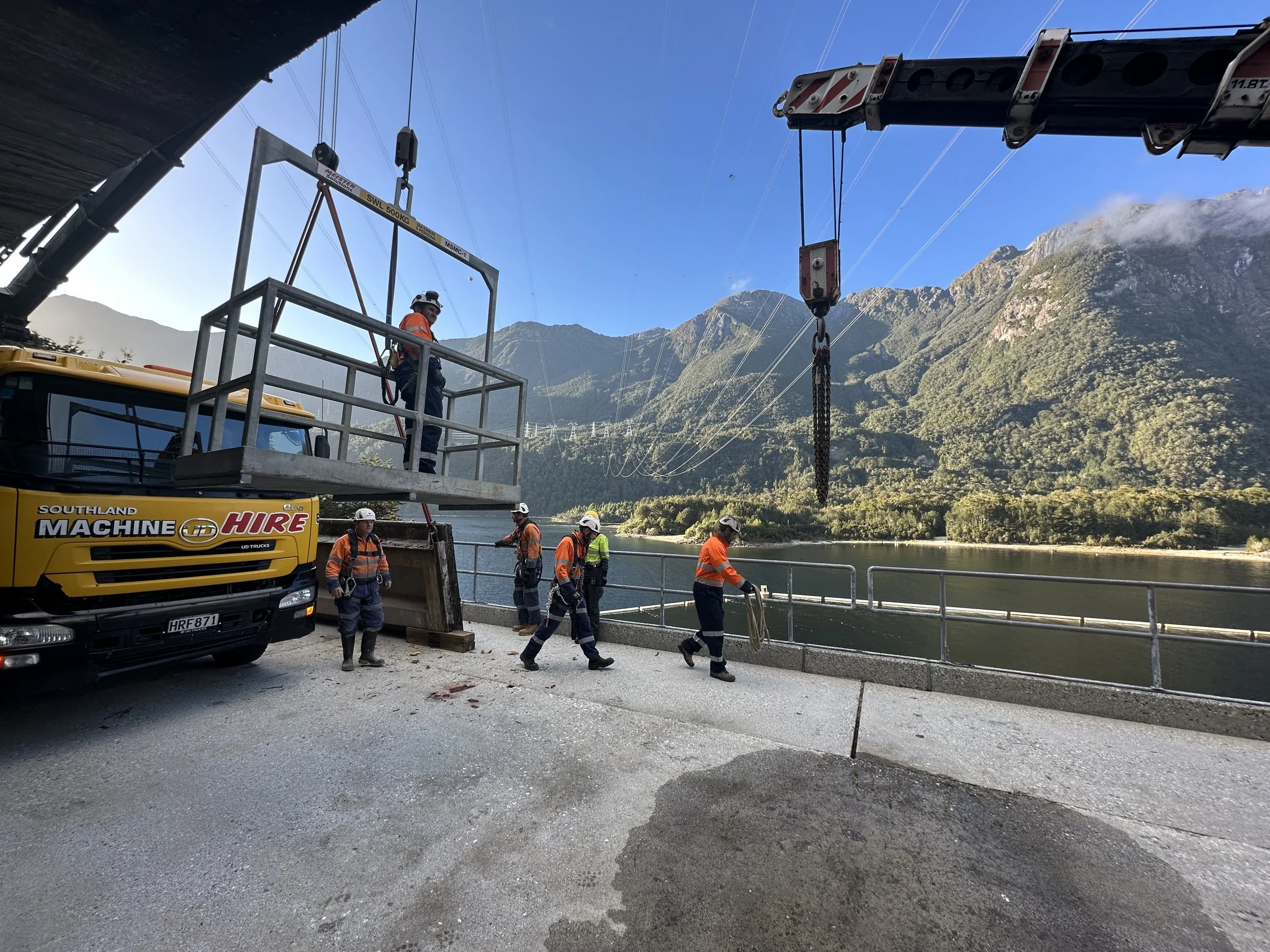Workers in orange safety gear and helmets are working with a crane near a body of water, with mountain ranges in the background.