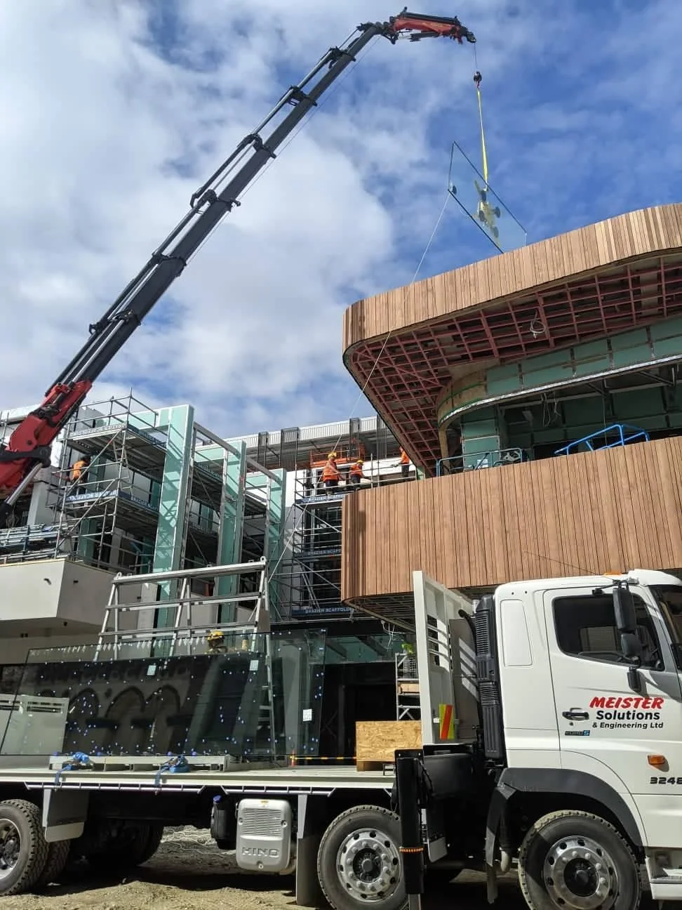 Construction workers on scaffolding installing glass panels on a modern wooden building, with a crane lifting a glass sheet into place under a partly cloudy sky.
