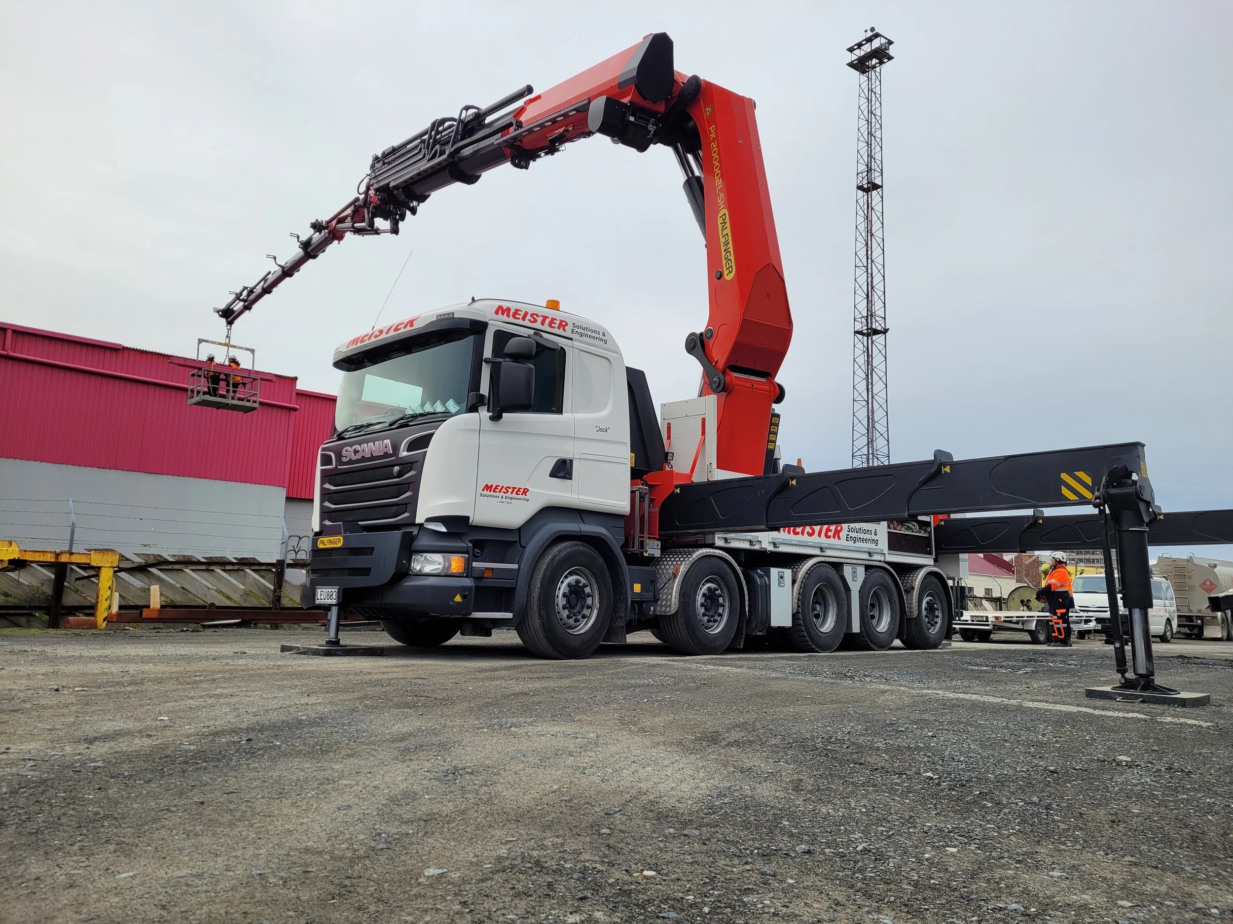 A large crane truck with a red and white cab, extending its boom overhead, at a construction site with workers in safety gear and a pink building in the background.