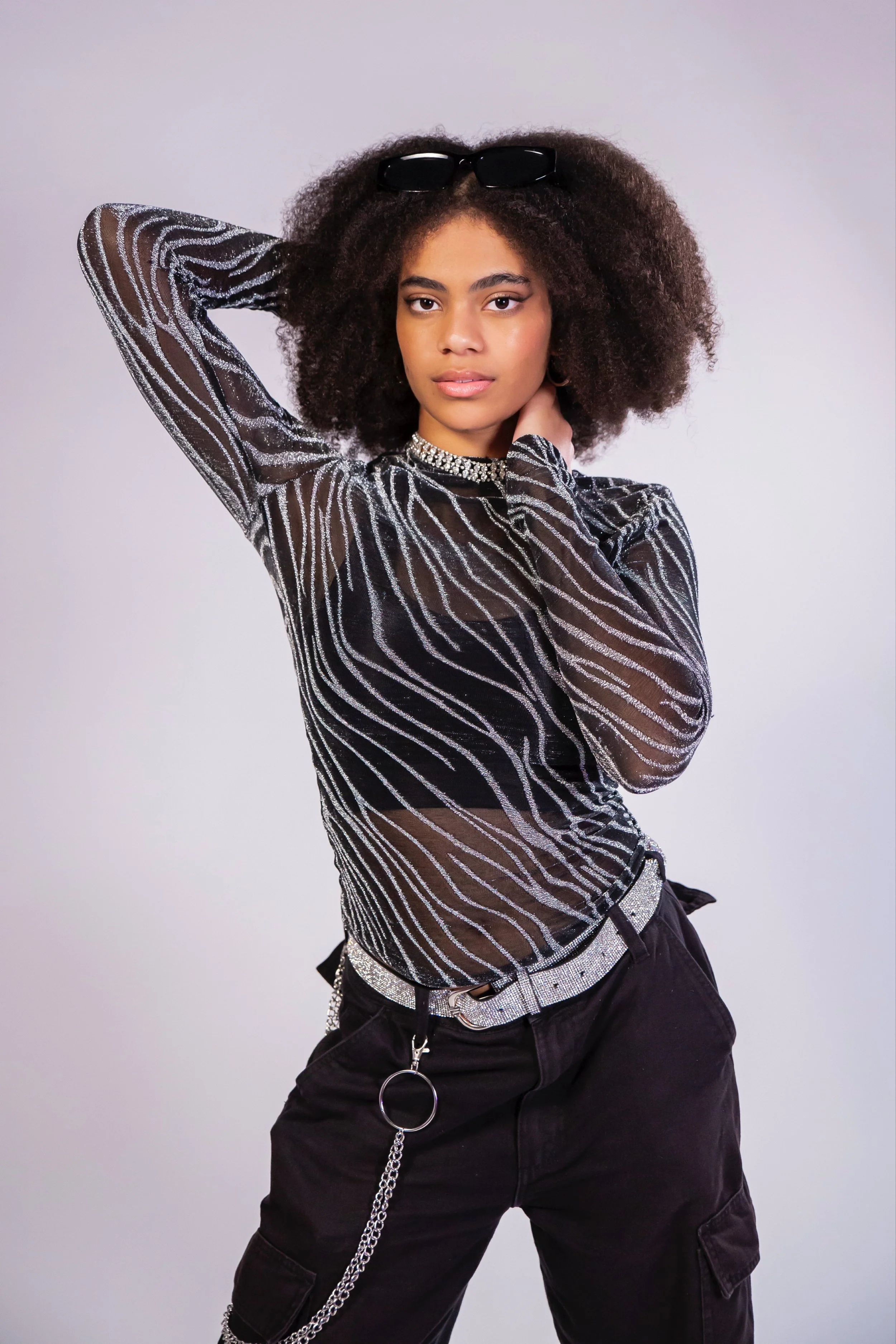 A young woman with curly hair wearing sunglasses on her head, a sparkly top, and black pants with a chain accessory, posing with one hand behind her head and the other near her neck.