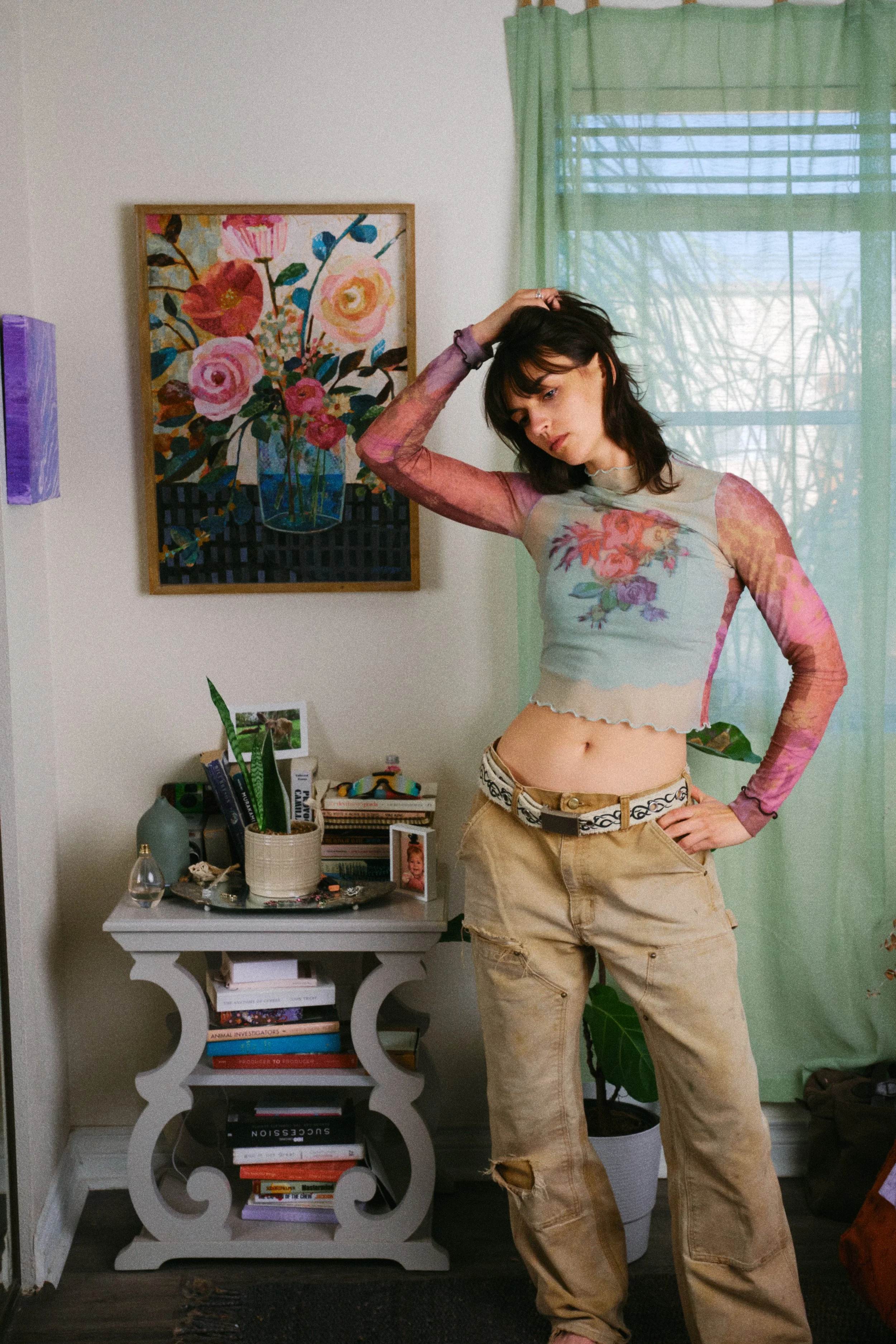 A woman with shoulder-length dark hair, wearing a semi-sheer pastel-colored floral long-sleeve top and beige pants, standing in a room with one hand resting on her head and the other on her hip, with a colorful floral painting on the wall, a green cu