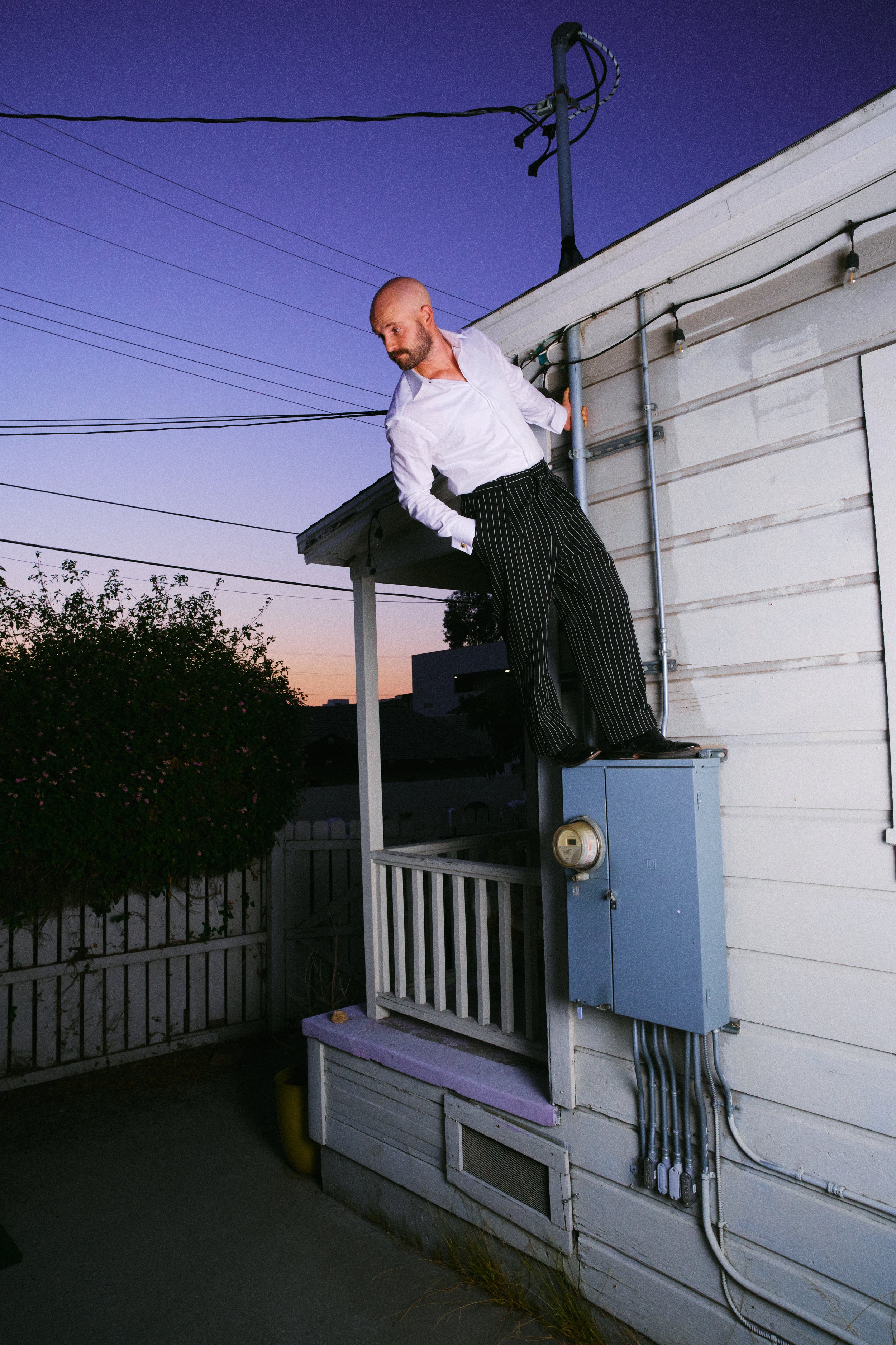 A man dressed in a white shirt and black pinstripe pants standing on an electrical box outside a house at dusk.