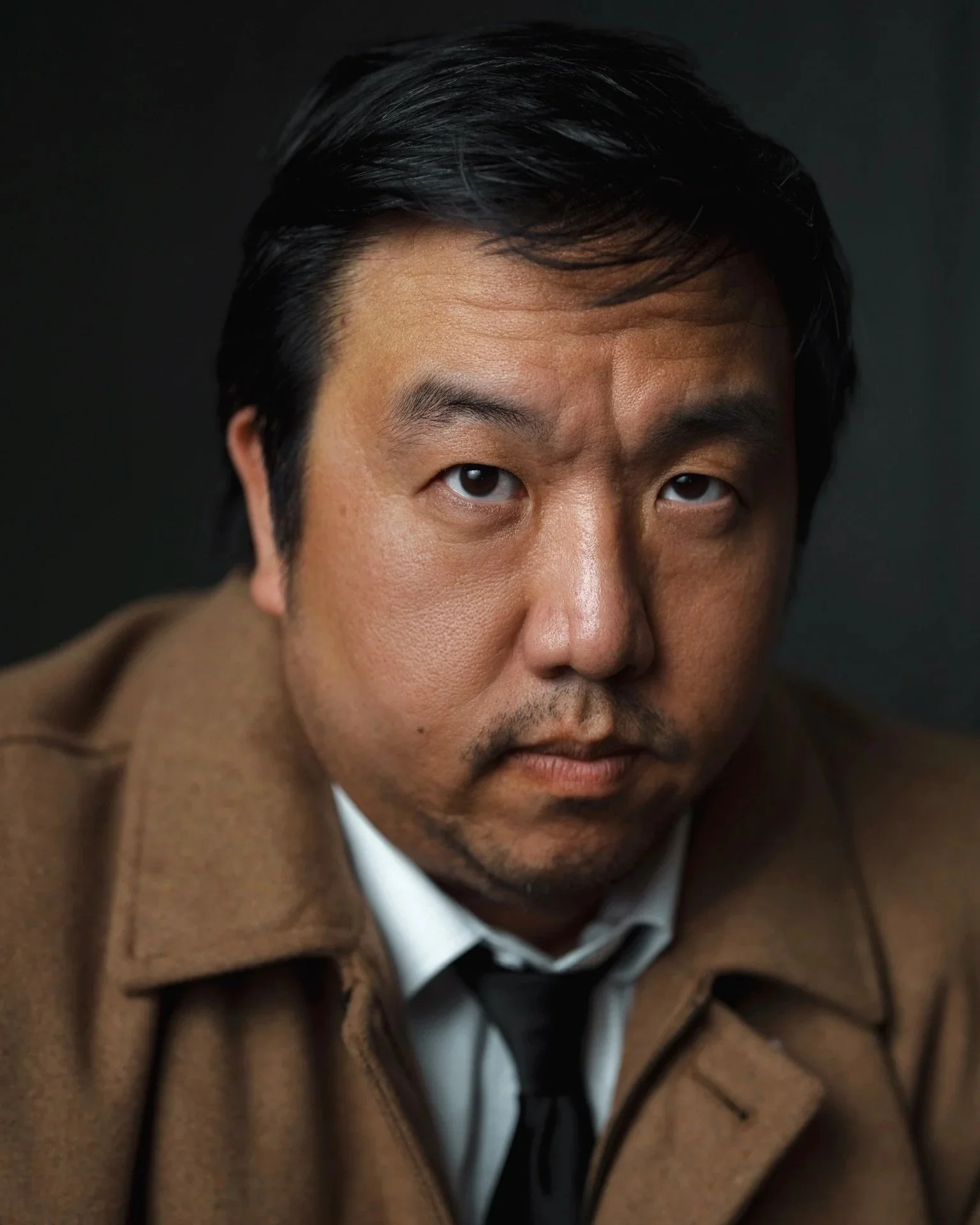 Close-up portrait of an Asian man with black hair, dressed in a tan coat, white shirt, and black tie, looking directly at the camera with a serious expression.