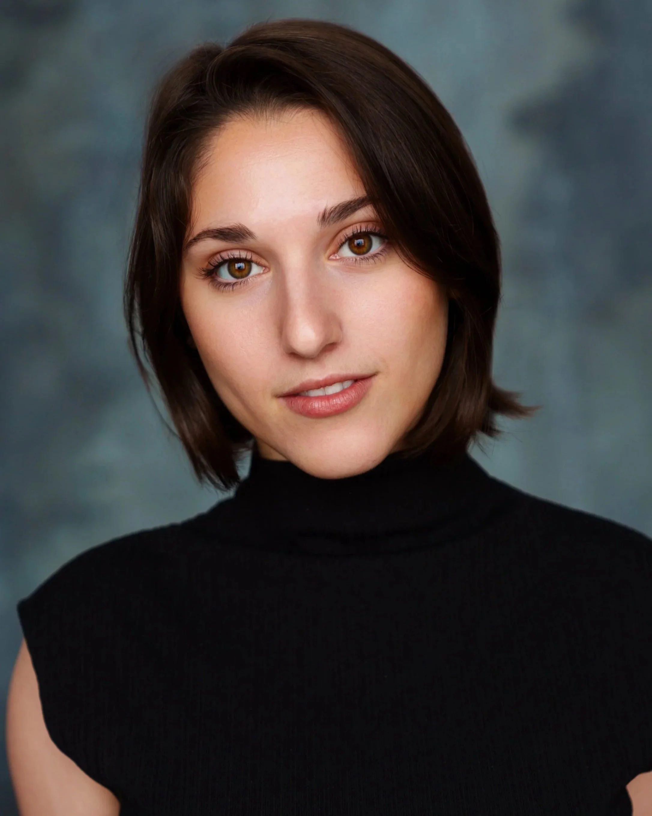 A woman with short brown hair and hazel eyes wearing a black turtleneck, looking at the camera, with a blurred blue and gray background.