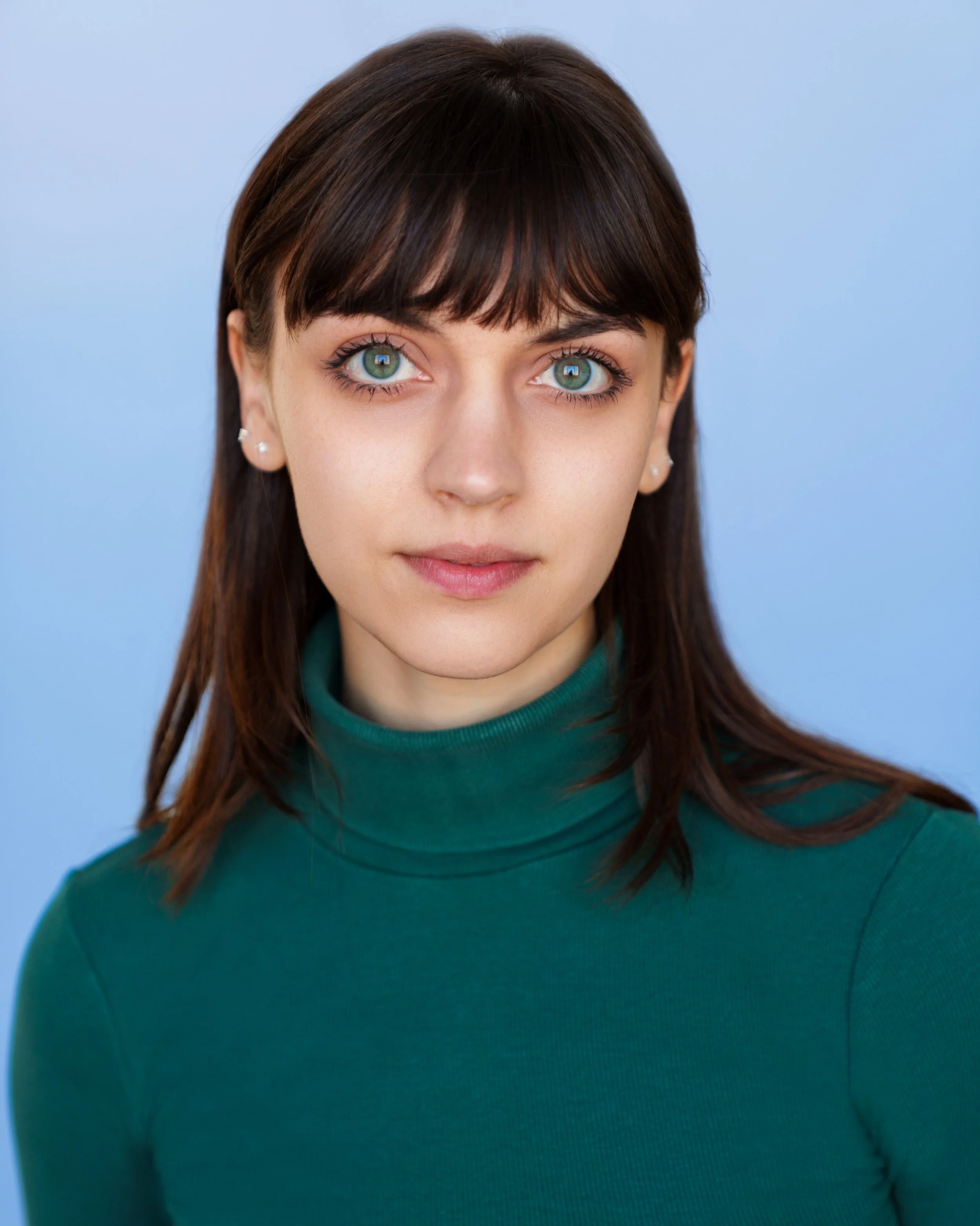 Portrait of a young woman with blue eyes and dark brown hair, wearing a green turtleneck sweater against a light blue background.