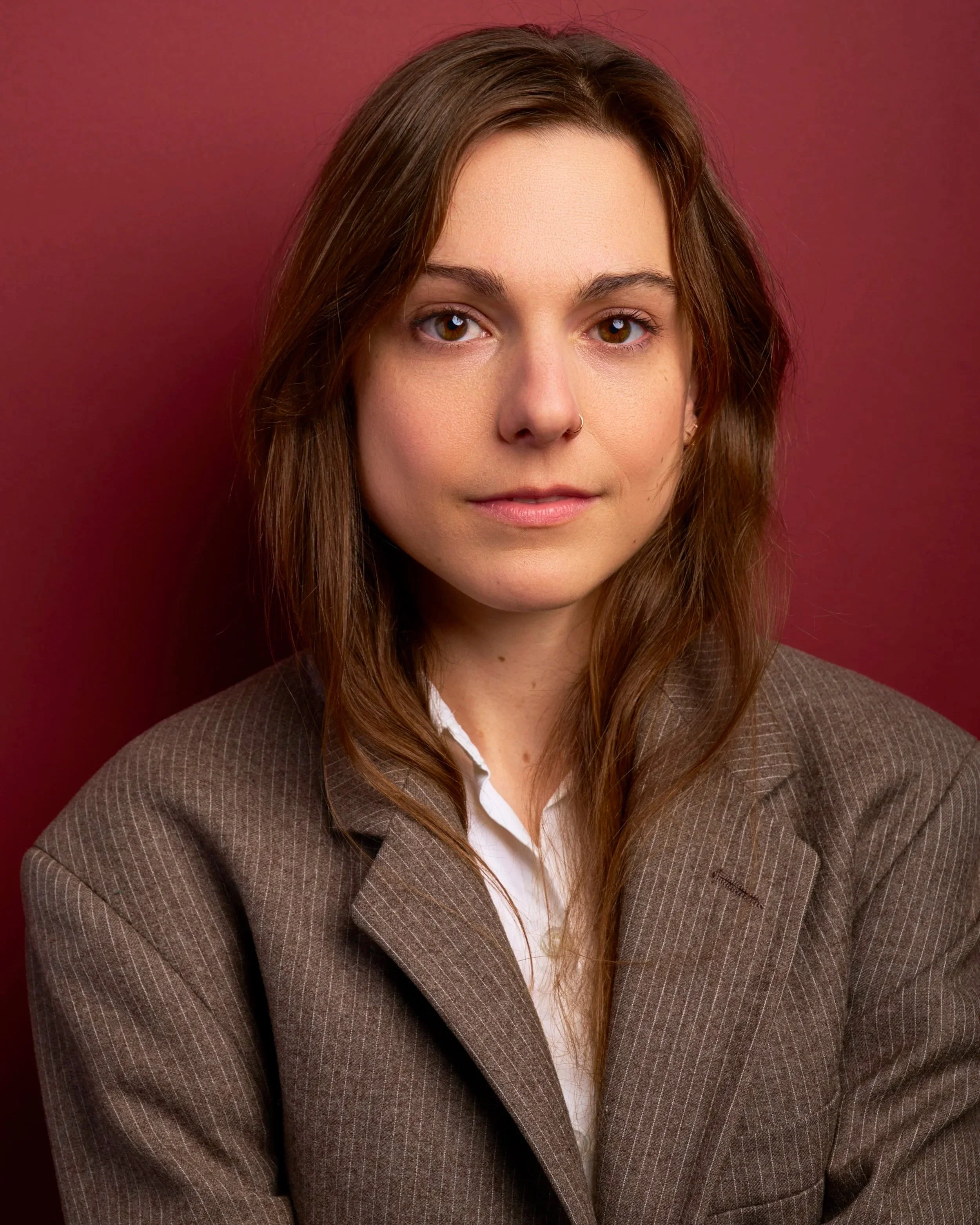 A young woman with brown hair and brown eyes, wearing a brown pinstriped blazer over a white shirt, posing against a reddish-brown background.