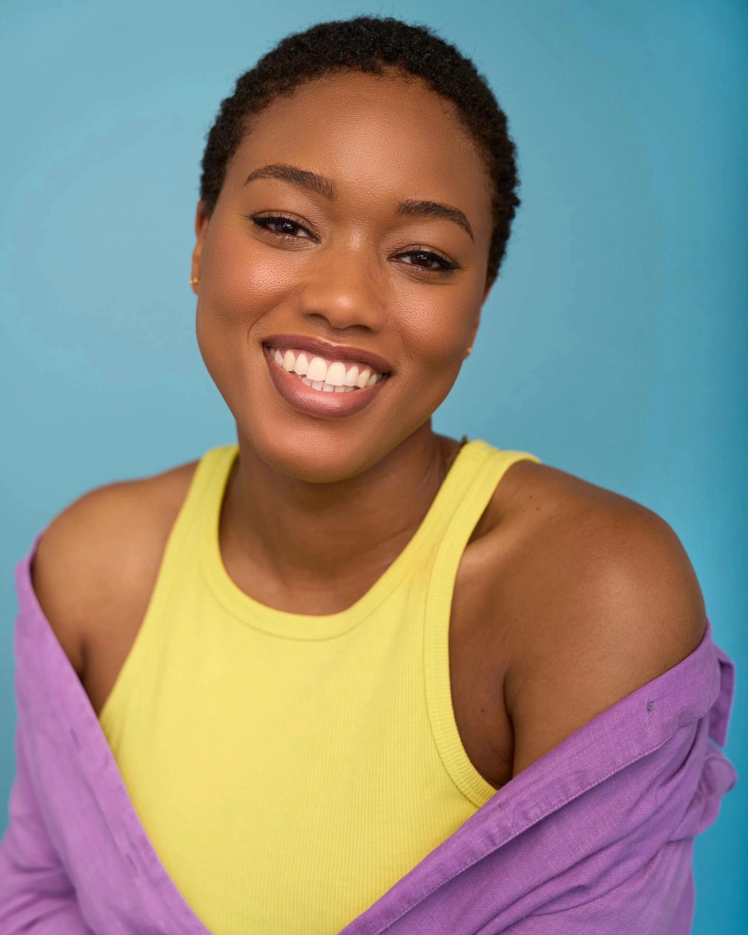 A smiling woman with short curly hair wearing a yellow tank top and a purple shirt draped over her shoulders, set against a blue background.