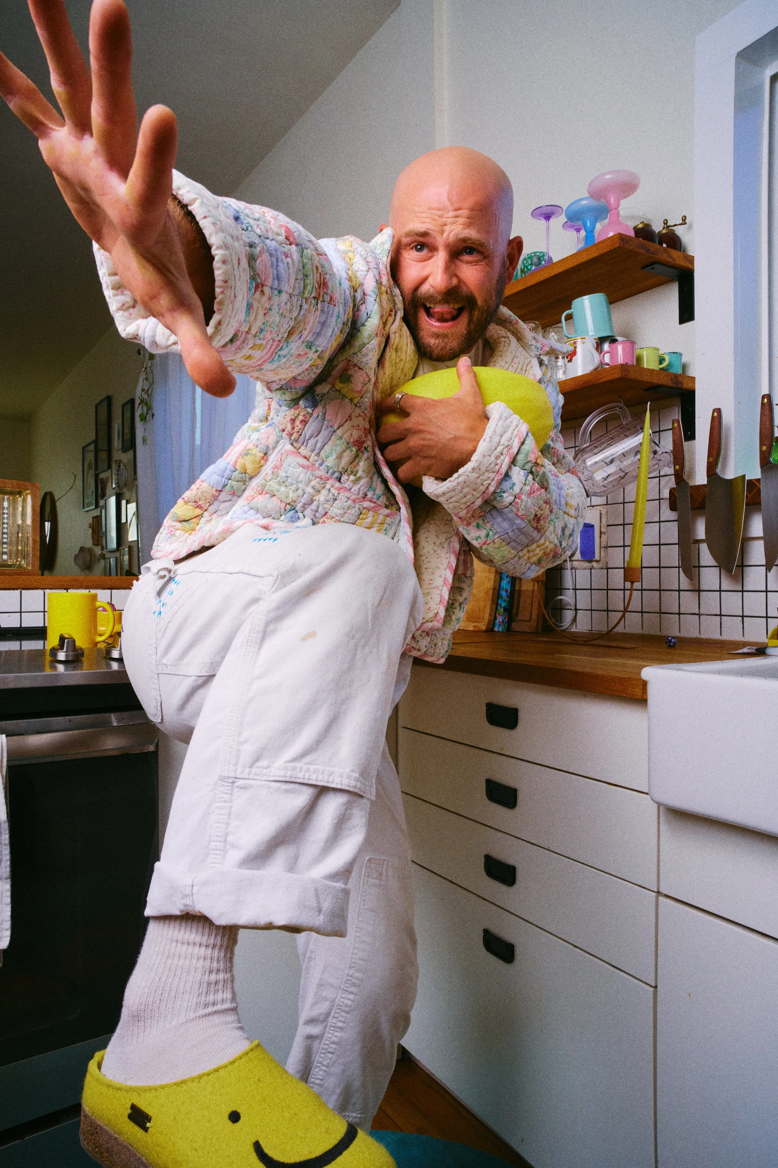 A man with a bald head and beard in a kitchen, reaching forward with an expressive face, clutching a yellow object, wearing a quilted jacket, white pants, and smiling yellow slippers.