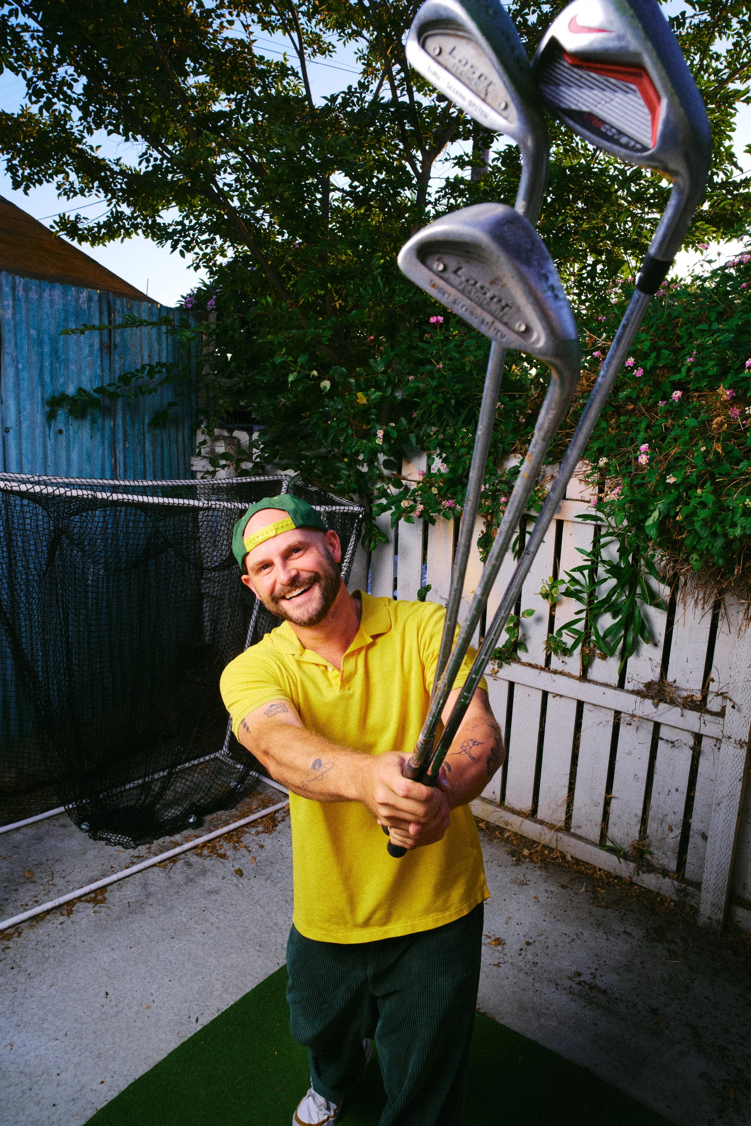A man in a yellow shirt and green cap smiling and holding golf clubs in an outdoor backyard setting.