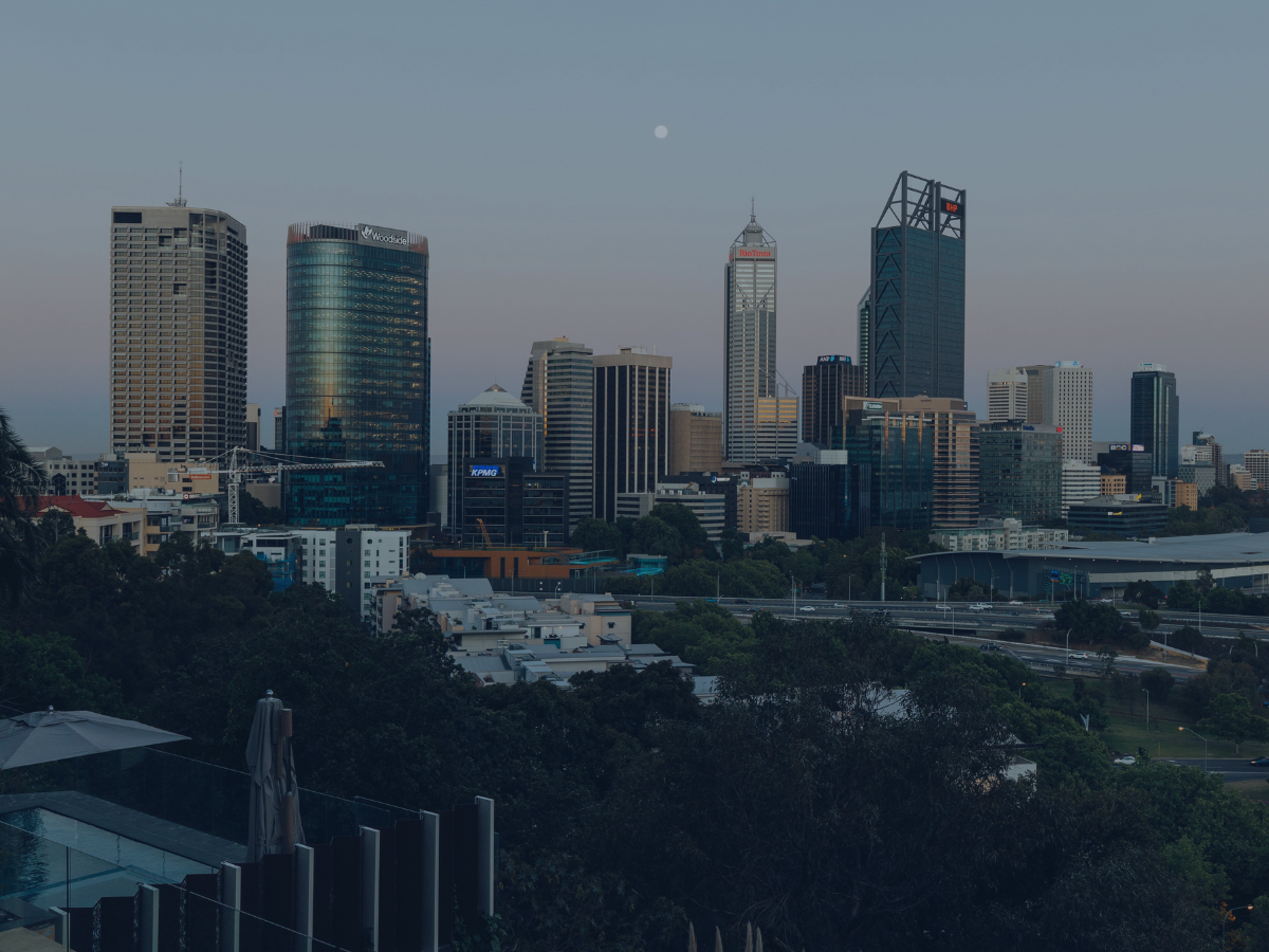 City skyline with tall modern skyscrapers under a clear evening sky, moon visible above the buildings, and some trees and rooftops in the foreground.