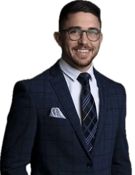 A young man with glasses and a beard wearing a dark blue plaid business suit, white dress shirt, and a matching angled plaid tie, smiling against a plain white background.