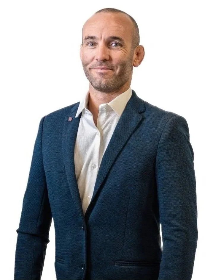 A man with a shaved head and a slight smile wearing a dark blue suit jacket and a white dress shirt, standing against a white background.
