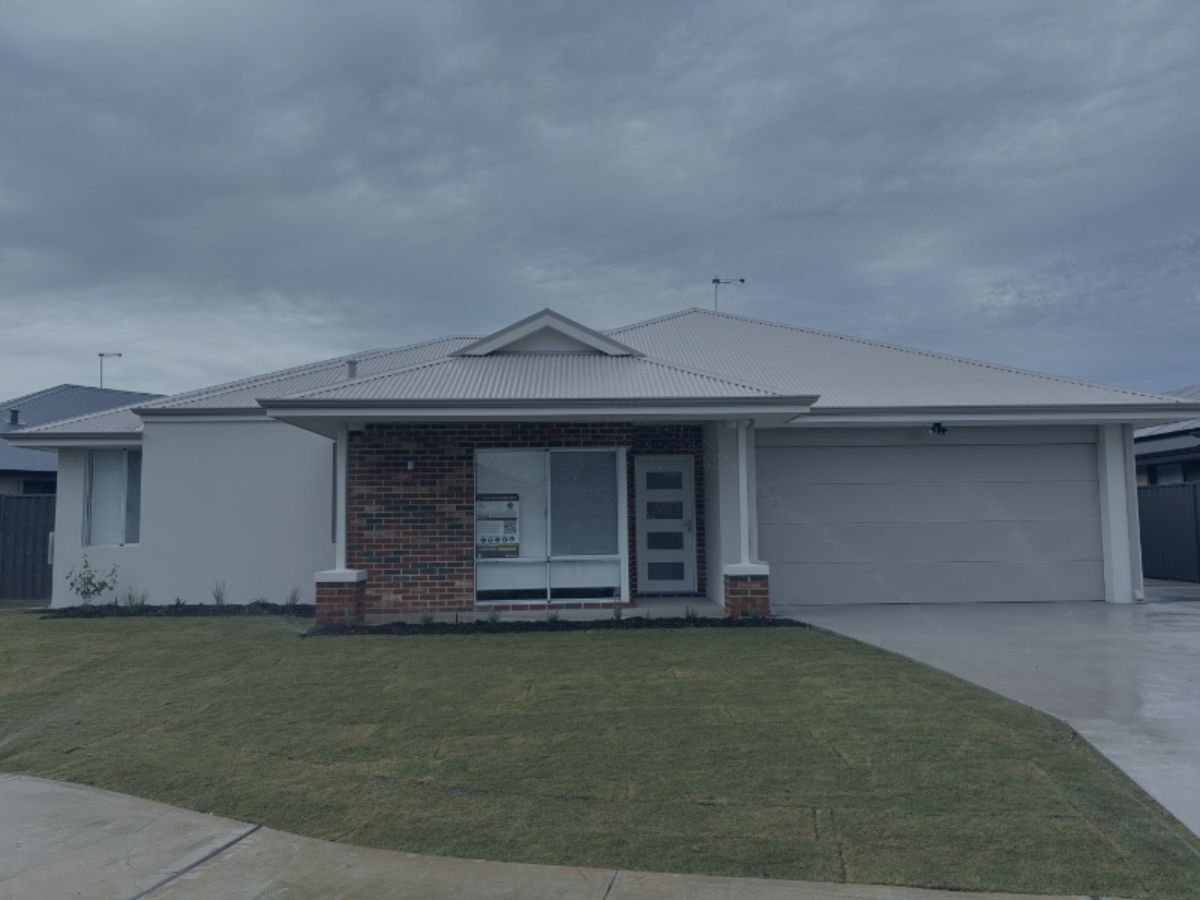 A modern house with a white exterior, brick accents around the front window, and a gray metal roof. The house has a driveway leading to a garage and a small front lawn under a cloudy sky.