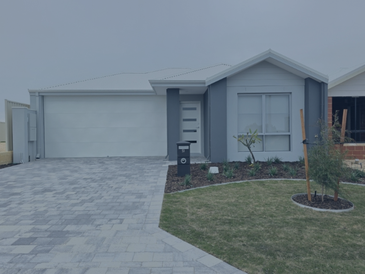 Front view of a modern single-story house with a gray garage door, a mailbox, a small garden with young plants, and a paved driveway.