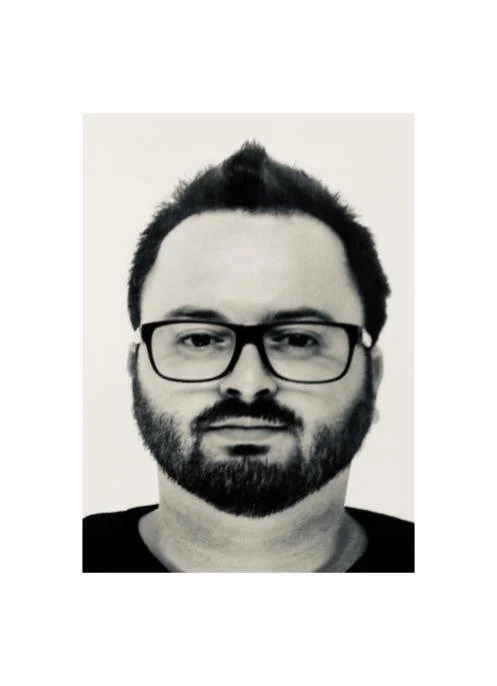 Black and white portrait of a man with glasses, a beard, and short dark hair, looking directly at the camera.