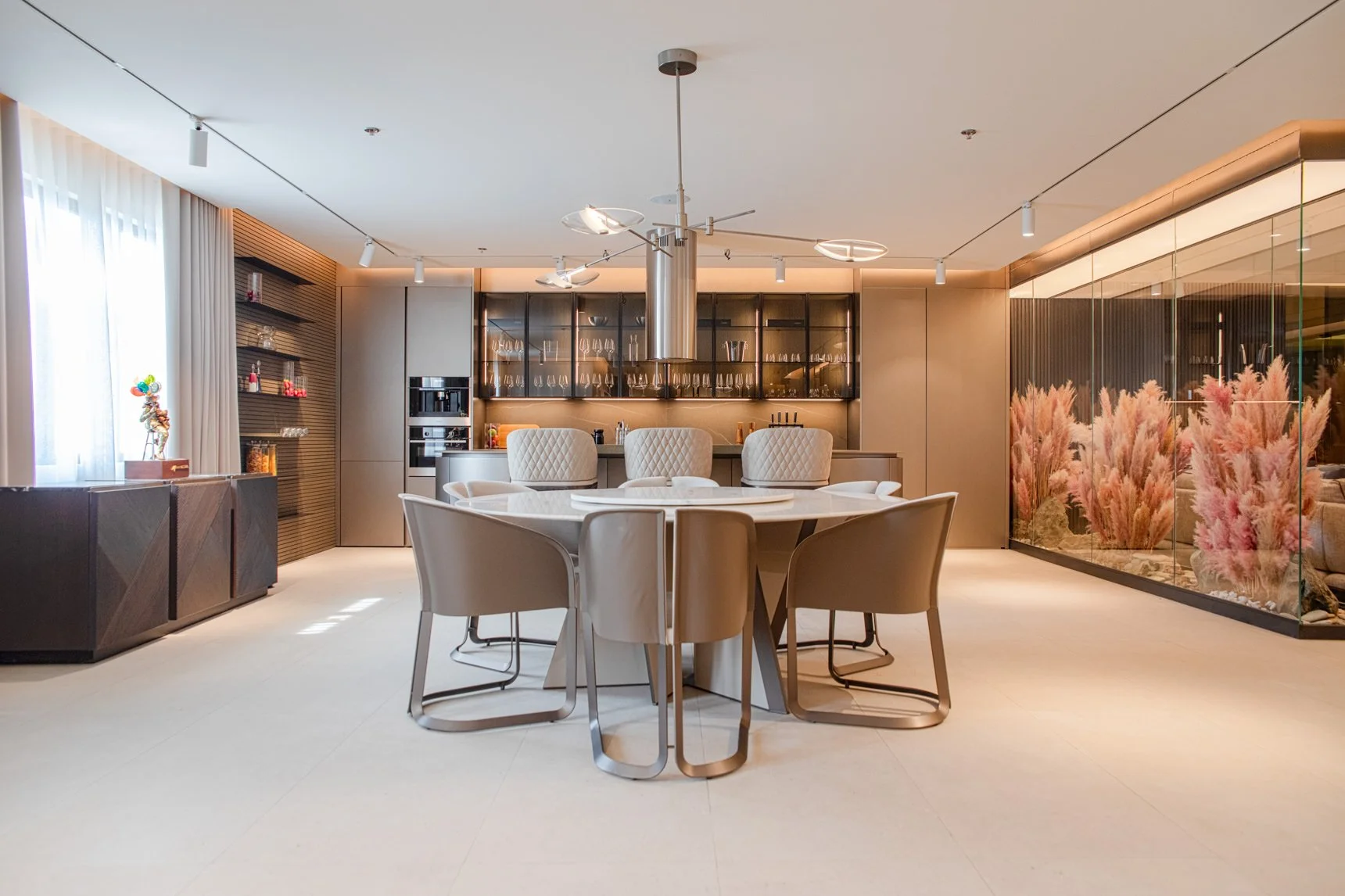 Modern dining area with a circular table surrounded by six beige chairs, set in a contemporary space with a glass wall of pink pampas grass and a kitchen in the background.
