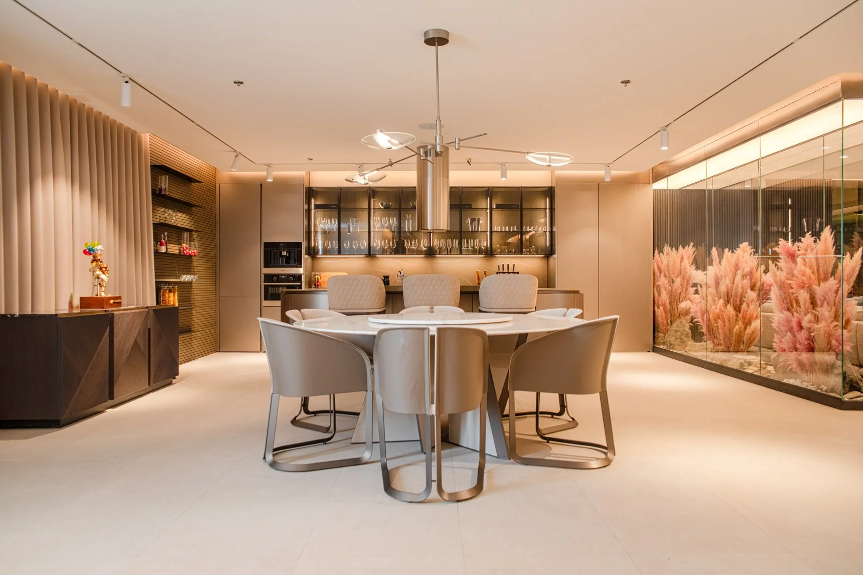 Modern dining room with round table, six beige chairs, stylish kitchen area, and large glass wall with pink ornamental plants.