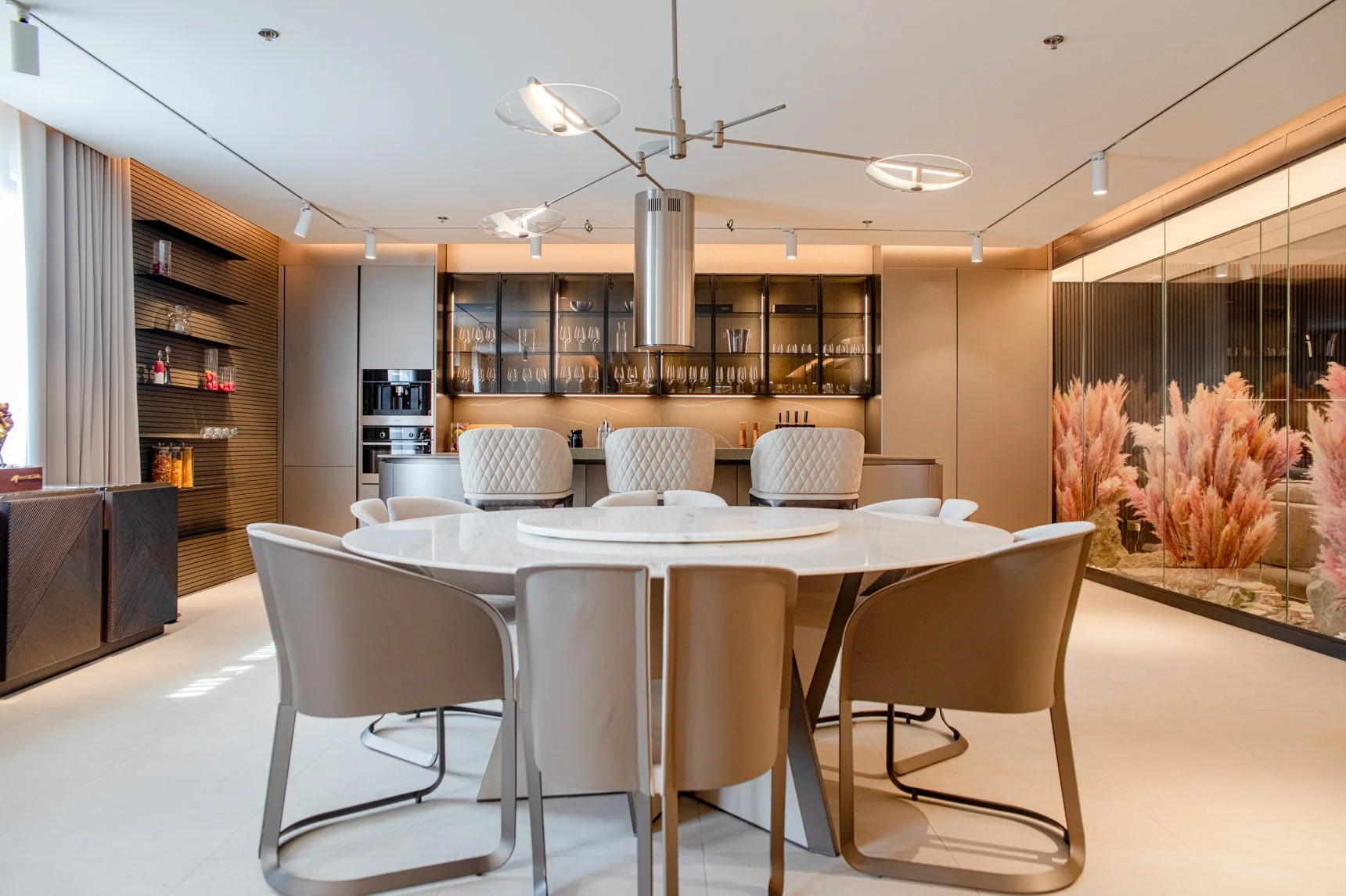 Modern dining room with a round marble table, eight beige chairs, a bar area with glassware and wine bottles, and decorative pink plants behind glass panels.