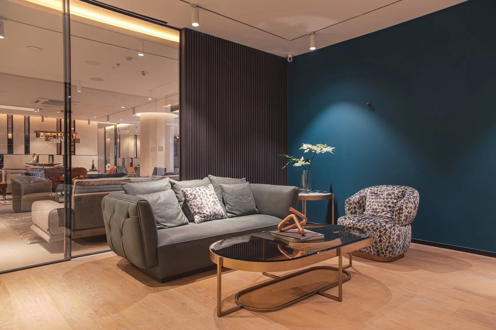 Modern living room with a gray sofa, a patterned accent chair, a black and gold coffee table, a side table with a tall vase of white flowers, and a dark teal accent wall.