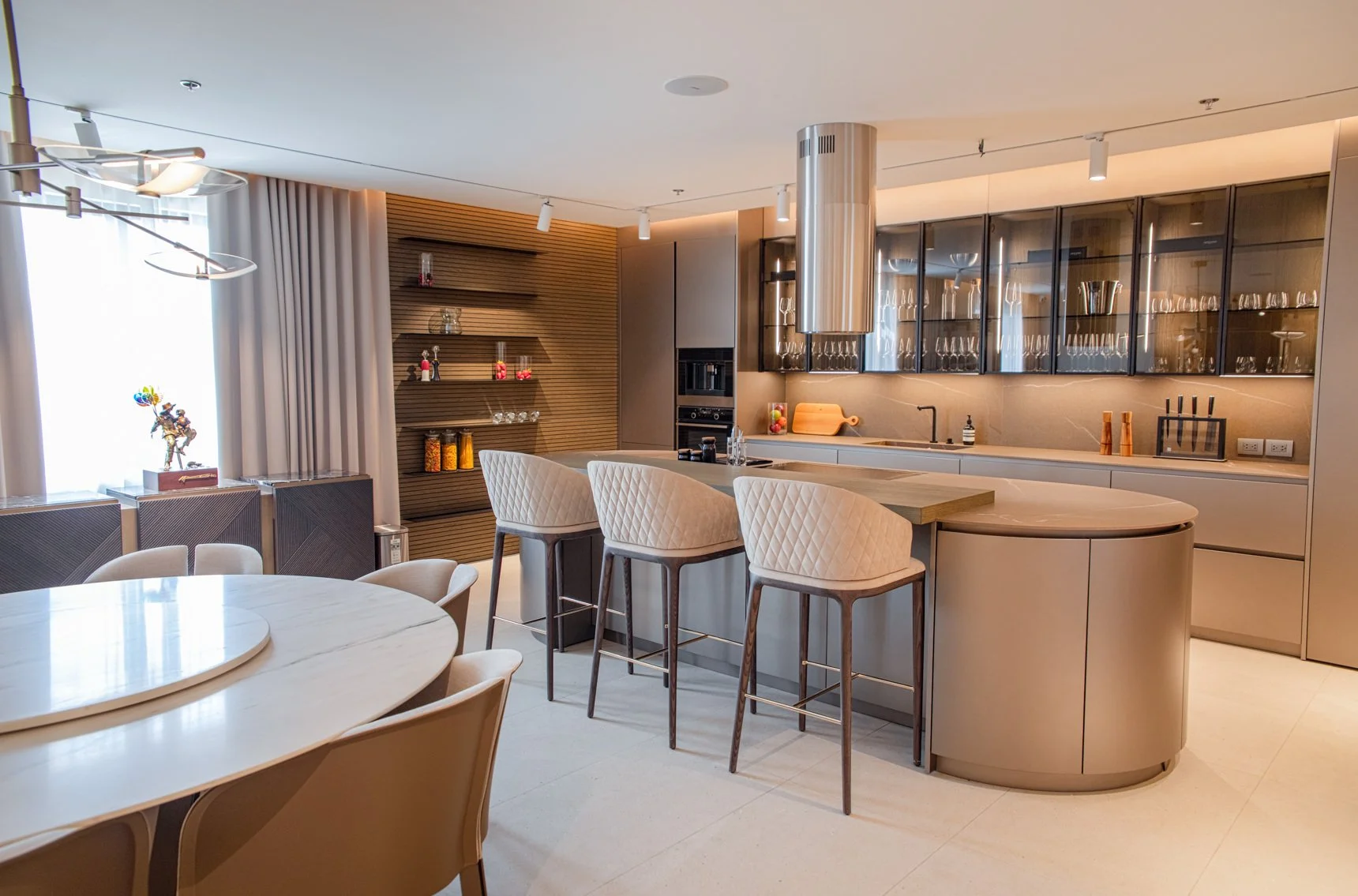 Modern kitchen and dining area with beige chairs around a table, bar stools at a curved kitchen island, glass-front cabinets, and decorative shelves, illuminated by natural and artificial light.
