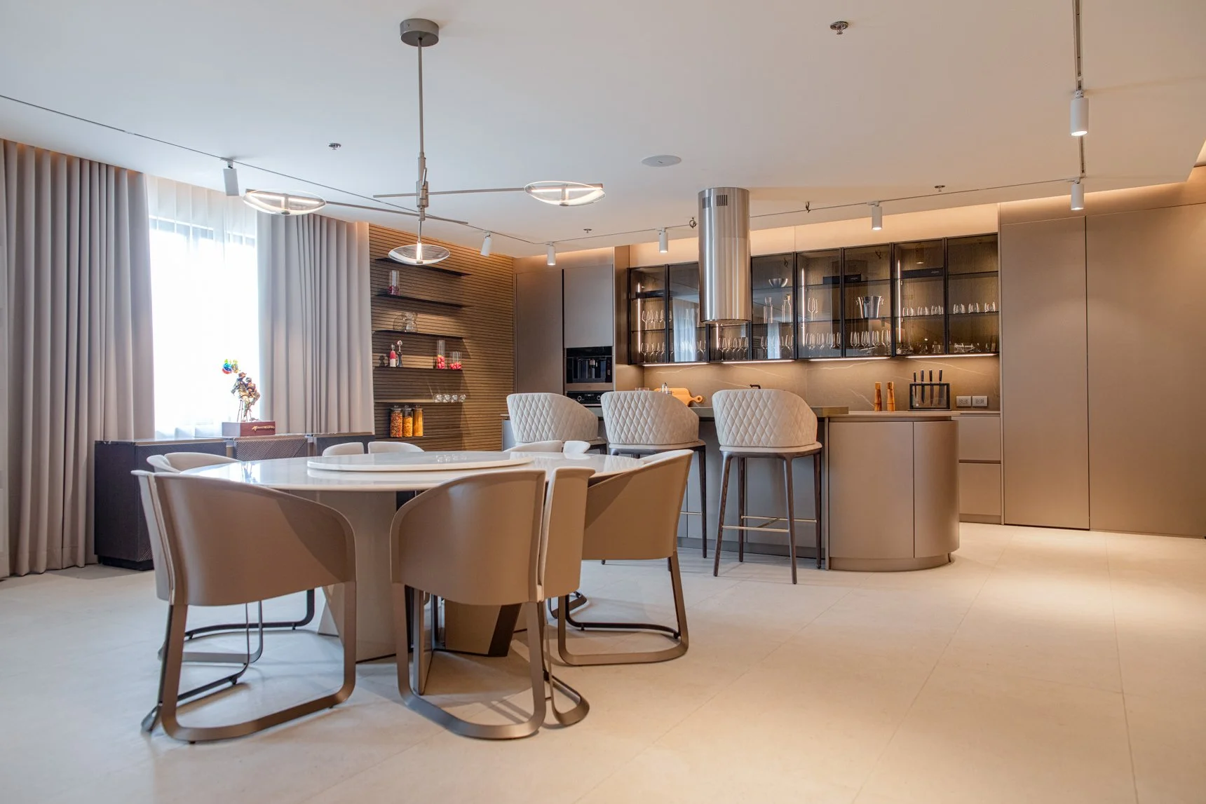 Modern kitchen and dining area with beige chairs around a round table, barstools at a curved kitchen counter, glass-front cabinets, and neutral decor.