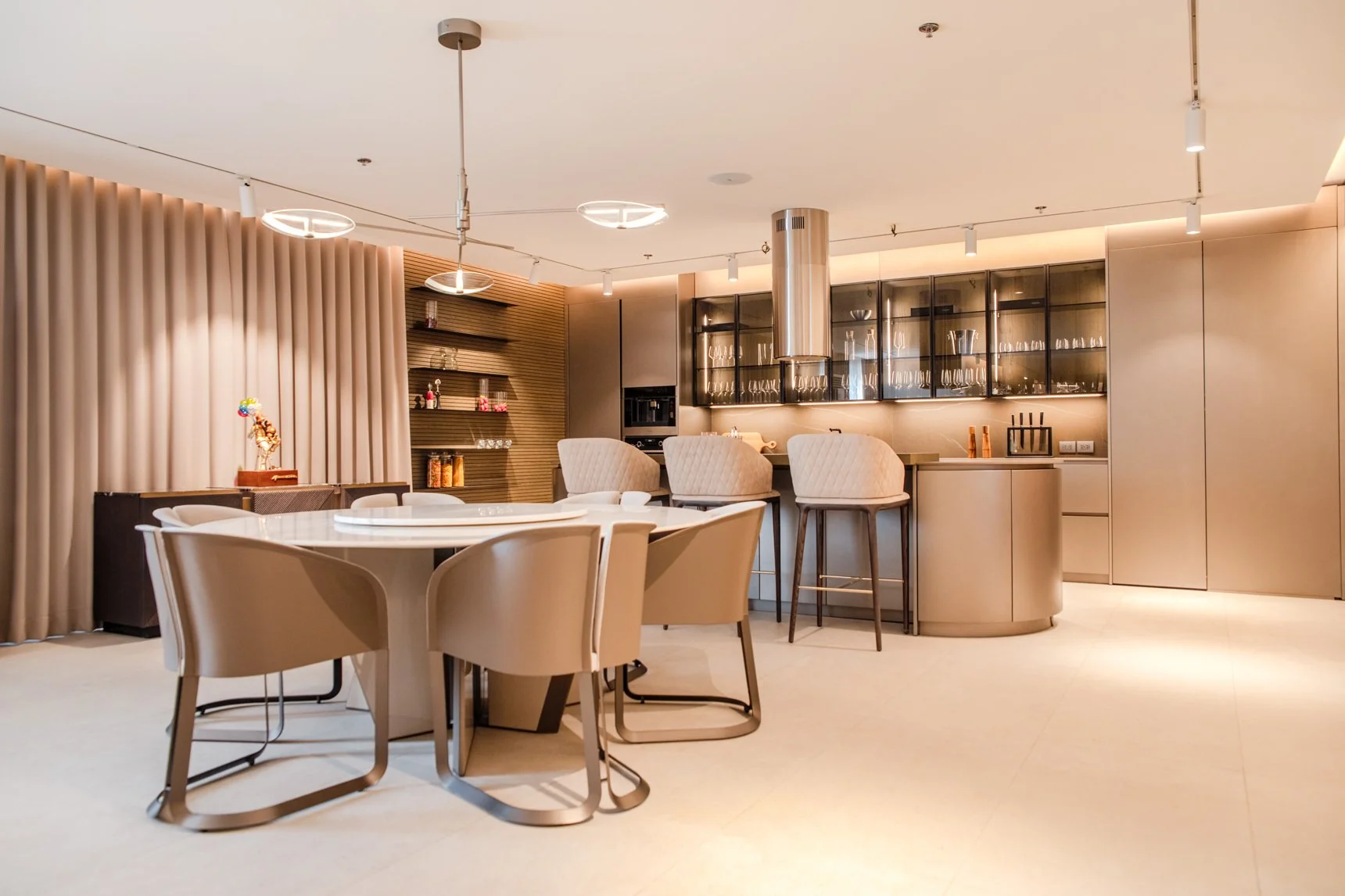 Modern kitchen and dining area with beige and metallic accents, a round table with chairs, bar stools at a breakfast bar, and built-in cabinets with glassware.