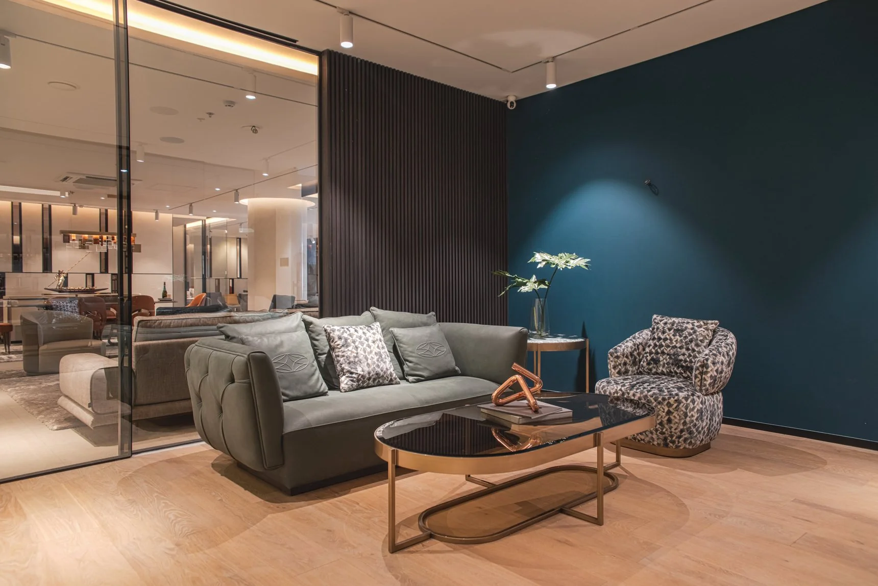 Modern living room with a dark green sofa, patterned armchair, black and gold coffee table, blue accent wall, and a side table with a vase of flowers.