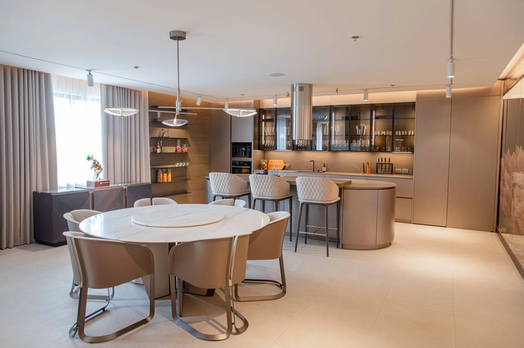 Modern kitchen and dining area with a round marble table, beige chairs, bar counter with high stools, and sleek cabinets with glass doors.