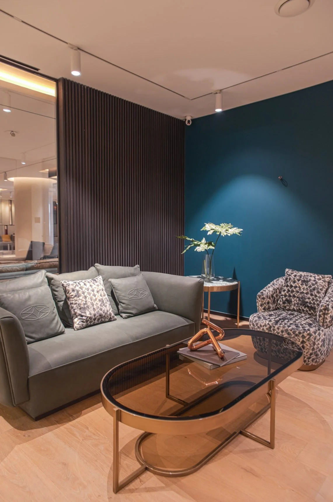 Modern living room with a green sofa, patterned armchair, glass coffee table, and a dark blue accent wall with a small table holding a plant, all in a stylish decor setting.