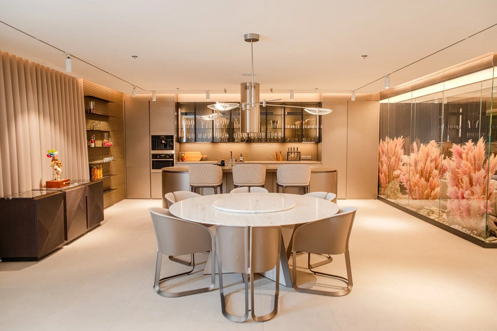 Modern kitchen and dining area with a round marble table, beige chairs, and a bar counter with four beige stools, decorated with pink ornamental grass in a glass wall.