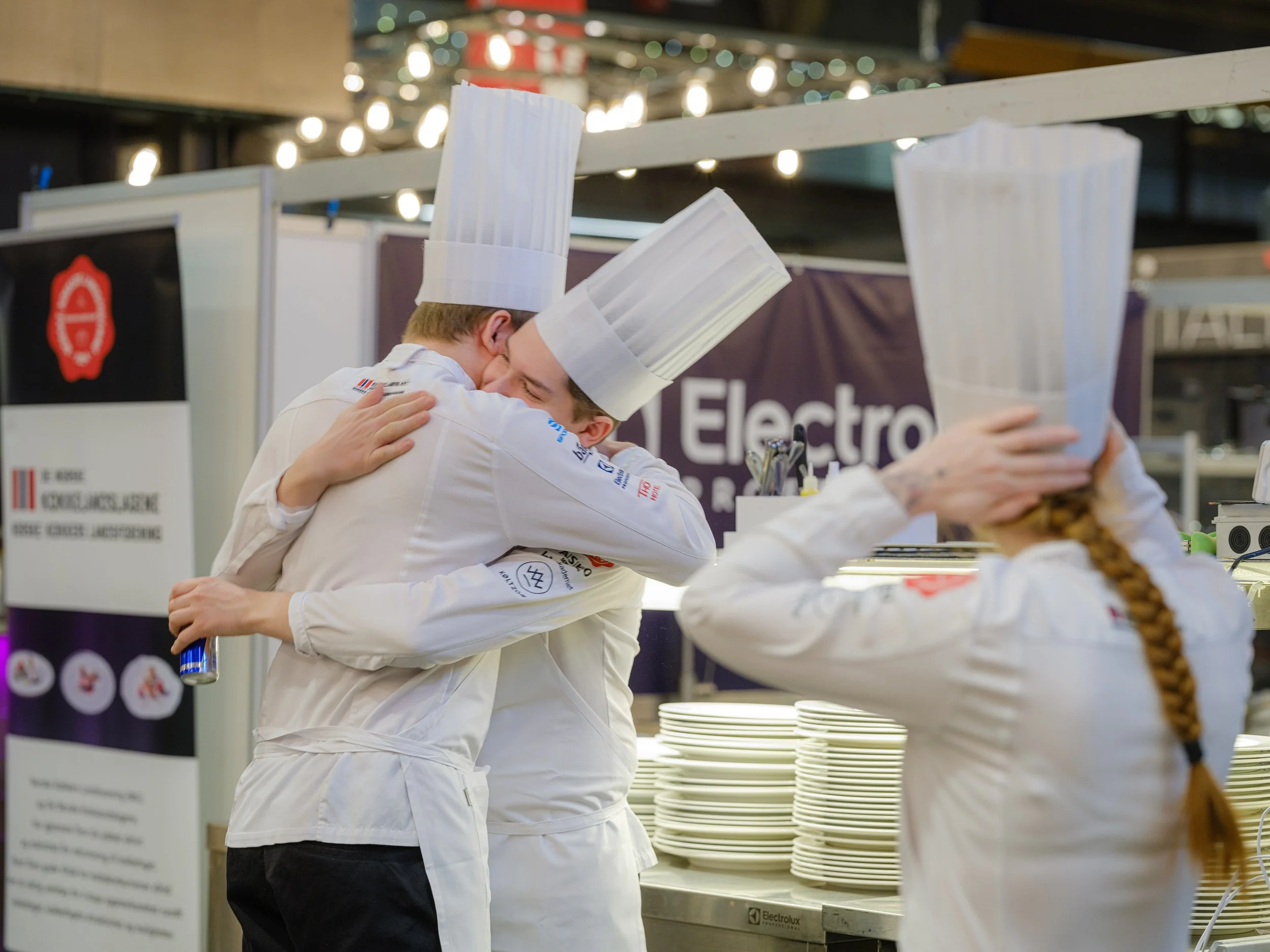 To kokker i hvite klær og kokkehatter klemmer hverandre i en varm omfavnelse på et kjøkken. En tredje kokk ser ut til å justere sin kokkehatt.