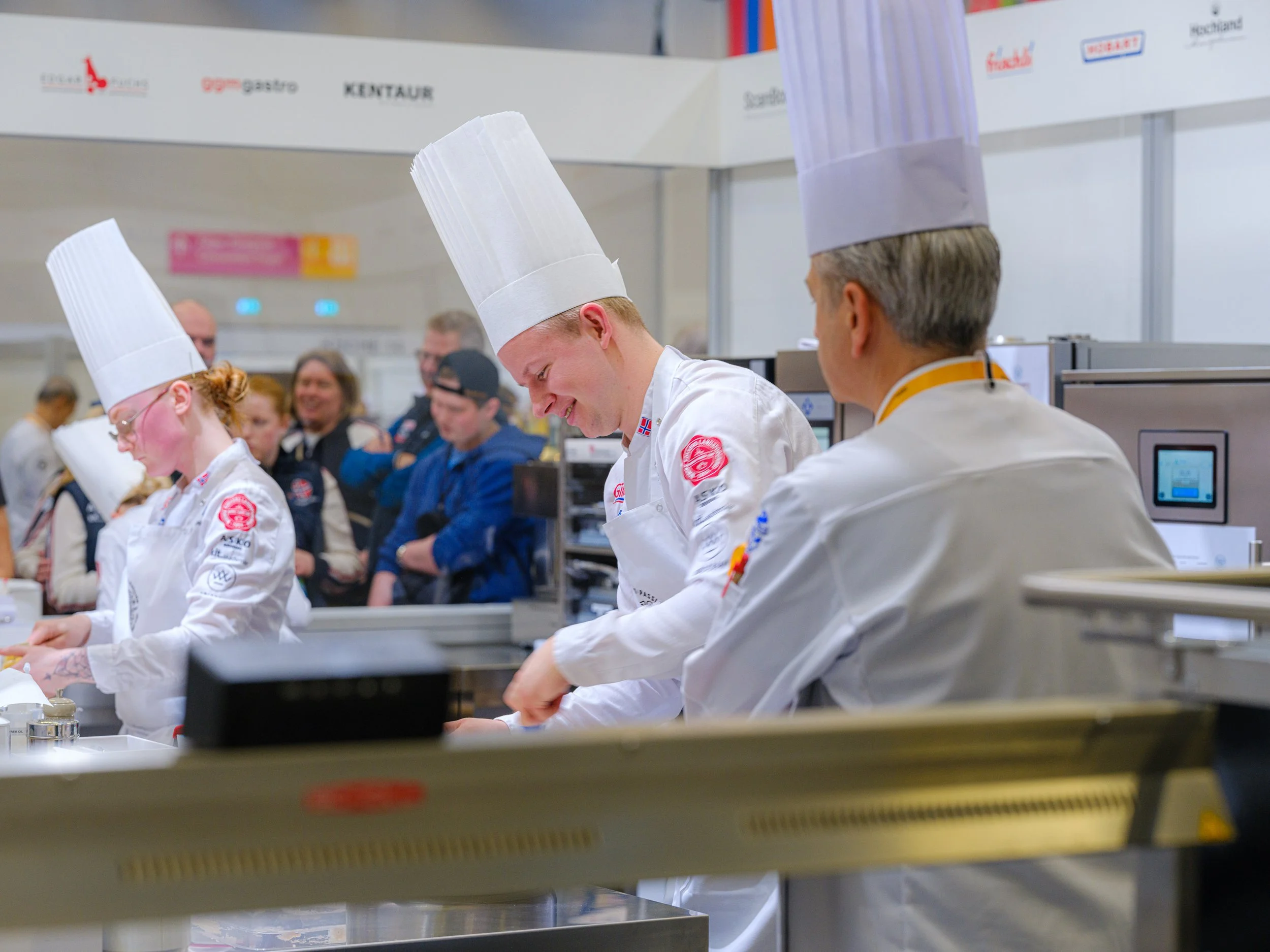 Kokkekamp med flere deltakere i hvite kokkeuniformer og høye kokkehatter, som smiler og arbeider i et profesjonelt kjøkken, med publikum i bakgrunnen.