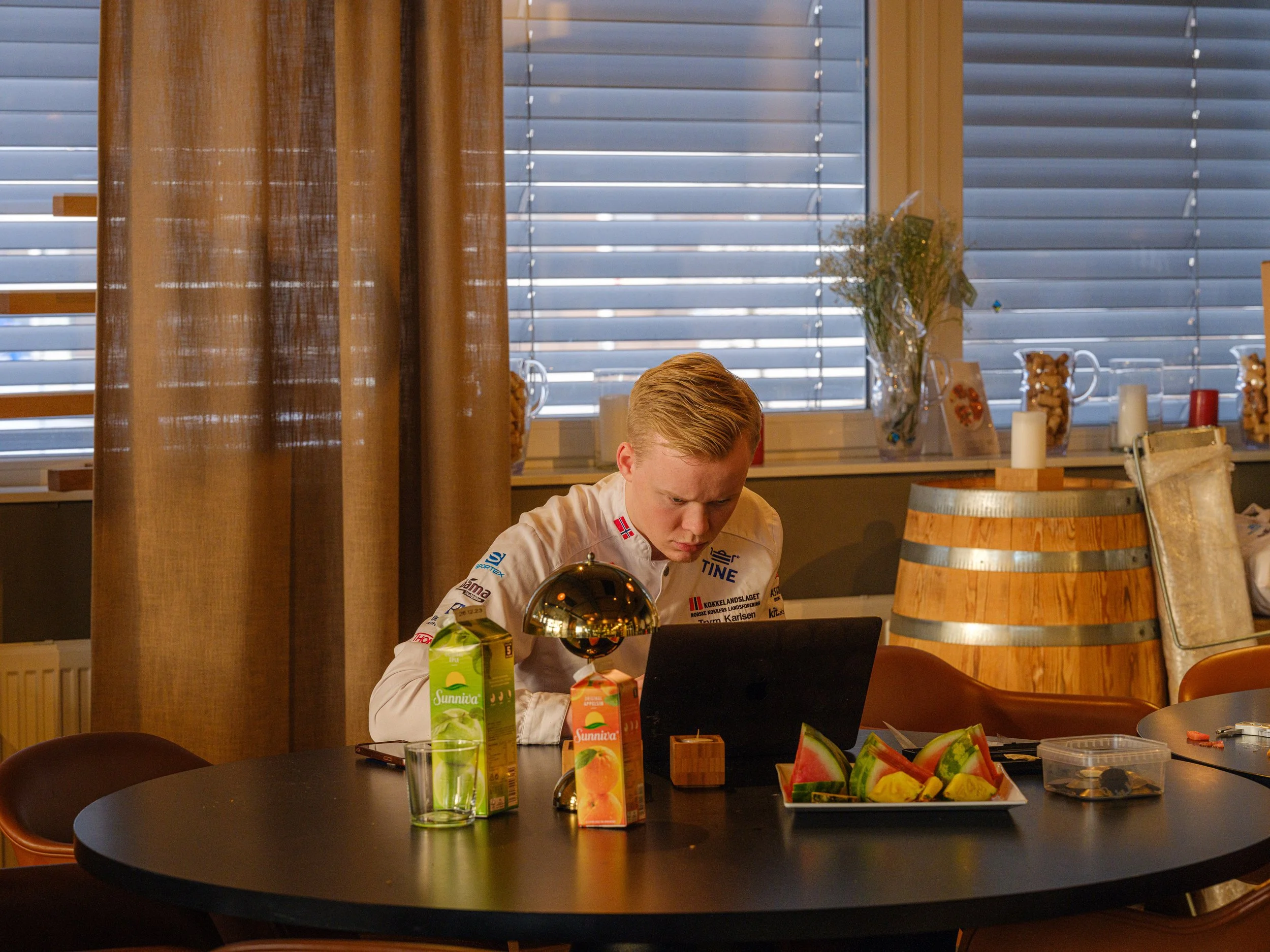 En ung mann som sitter ved et rundt bord med en bærbar datamaskin, flere juicetrefil og skiver vannmelon på bordet. Bakgrunnen viser et vindu med horisontale blinds og dekorative elementer på vinduskarmen.