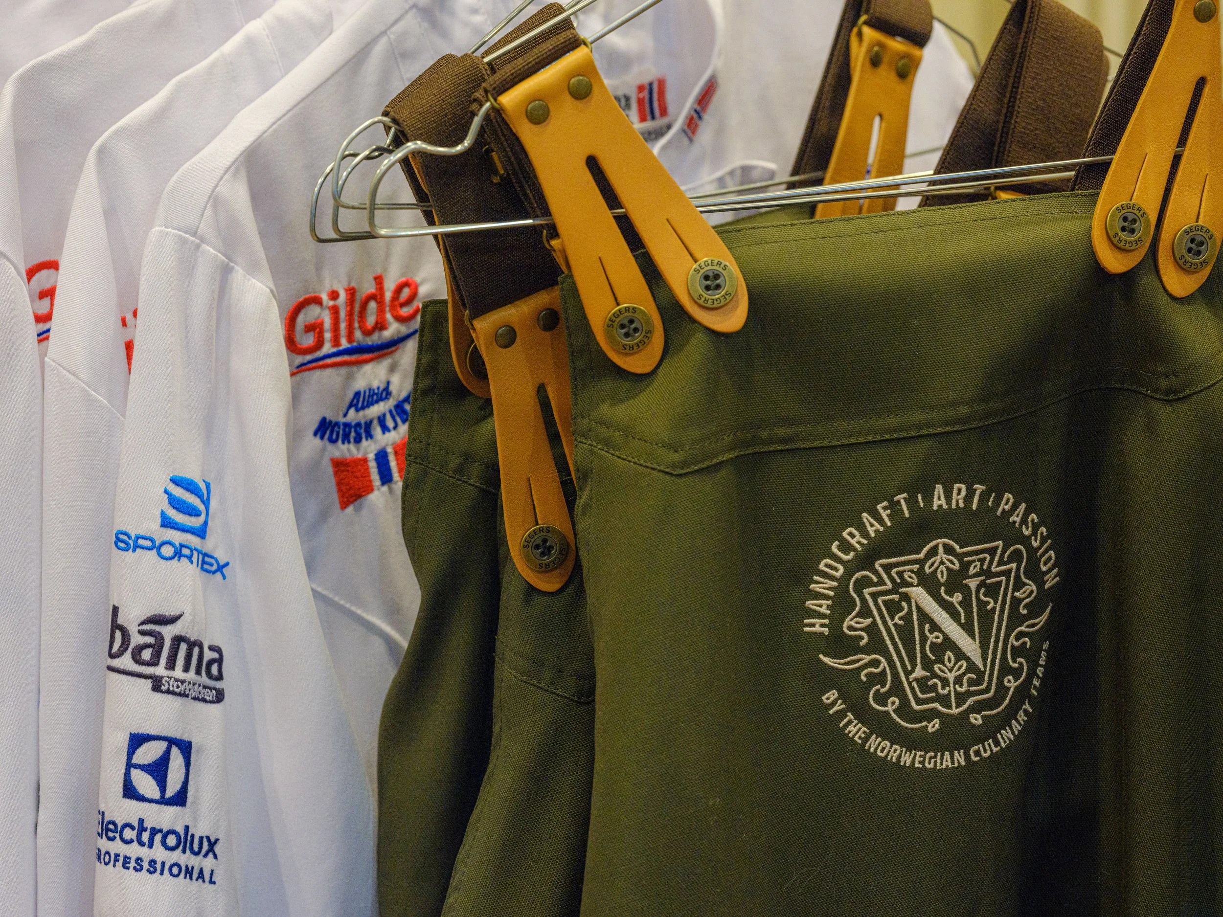 Klesplagg og verktøy hengende på kleshengere, inkludert en hvit uniform med logo og en grønn veske med tekst om norsk håndverkstradisjon.