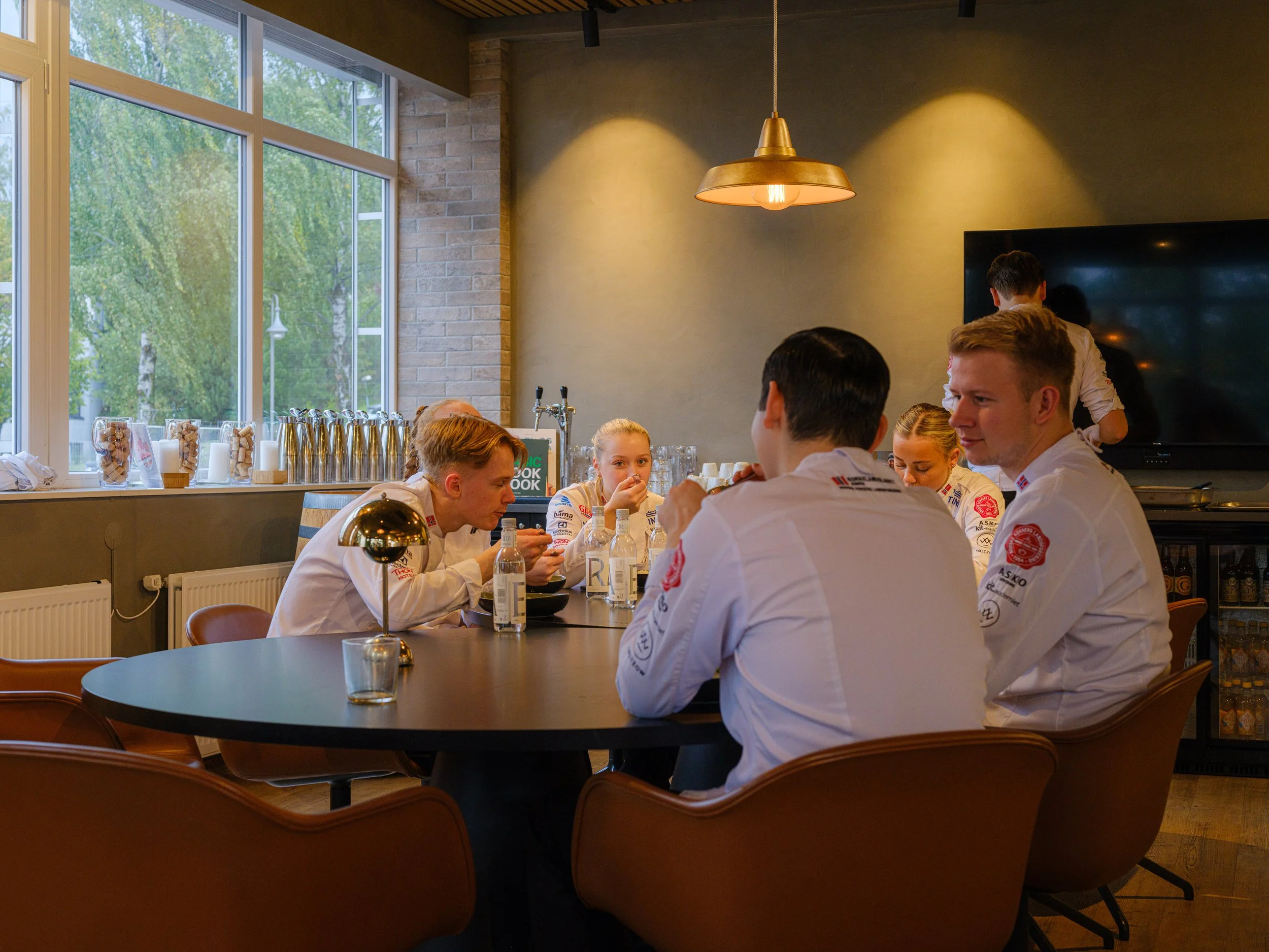 Fem personer i hvite kokkeuniformer sitter rundt et svart bord i en moderne restaurant eller pauserom, på ettermiddagen. Det er store vinduer i bakgrunnen som slipper inn naturlig lys, og det er ulike dryppende lamper i taket.