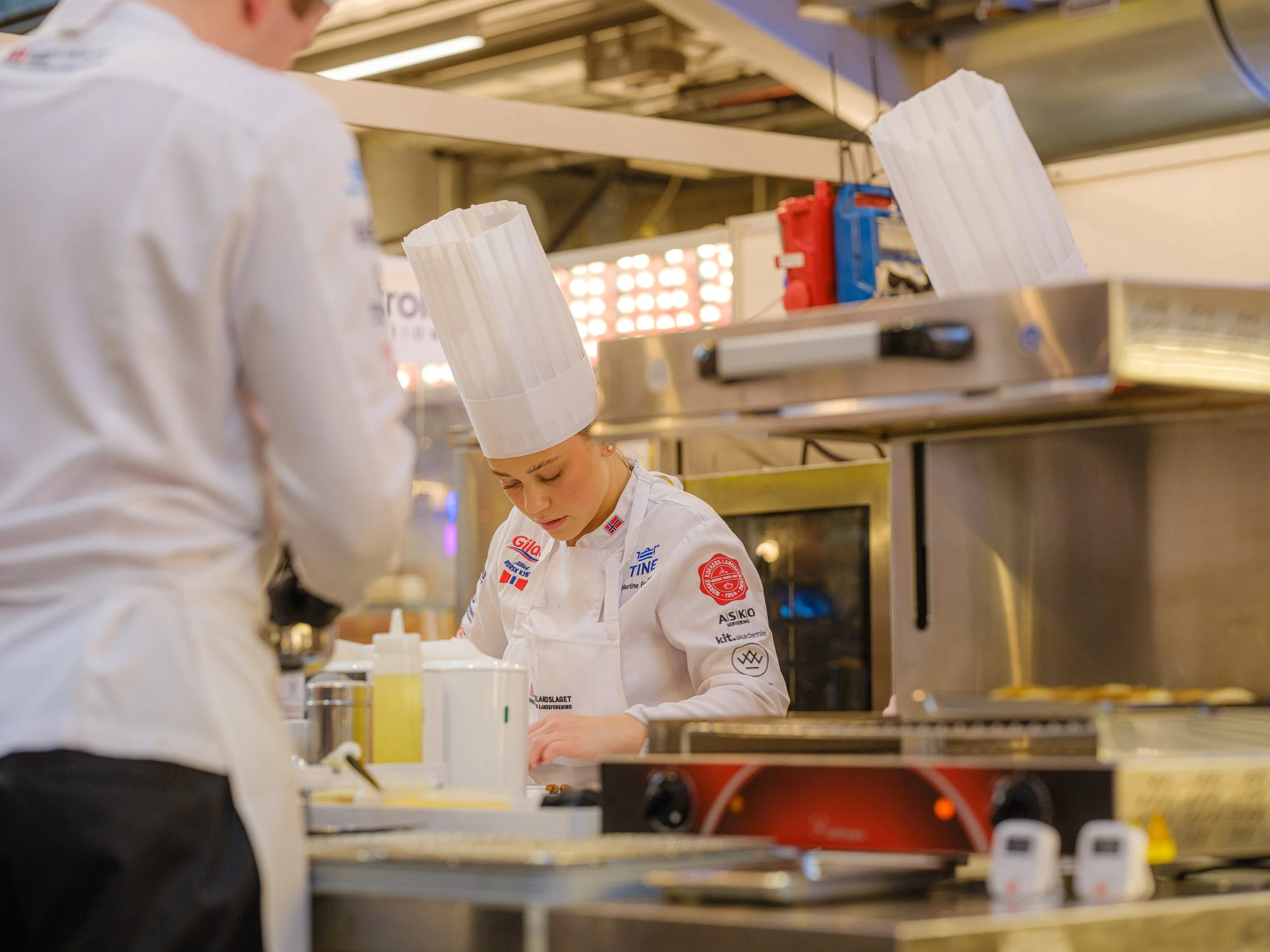 To kokker i hvite uniformer og kokkehatter jobber i et profesjonelt kjøkken.