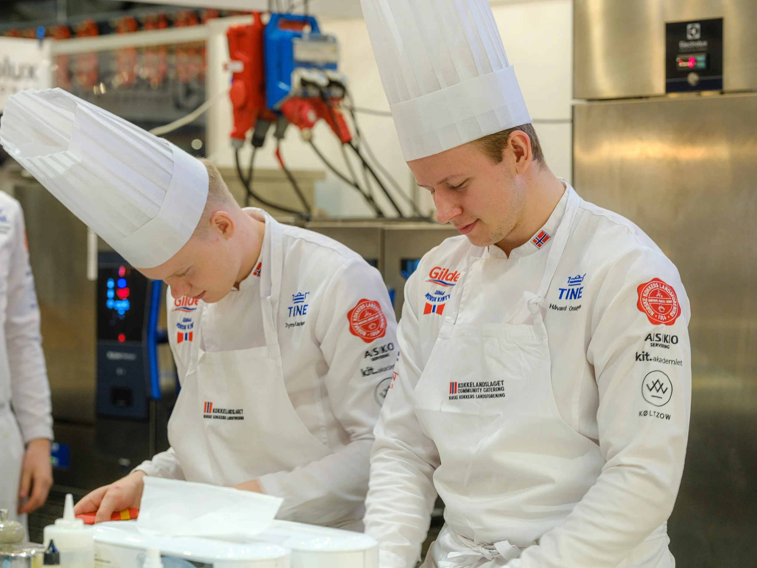 To mannlige kokker i hvite uniformer og kokkehatter jobber sammen i et kjøkken.