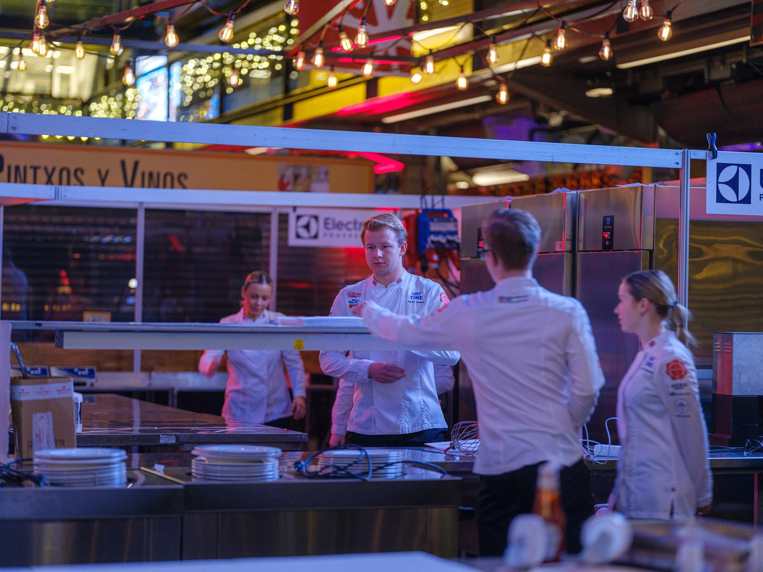 Fire personer i kokkeuniformer står i en kjøkkenlignende setting, på et sted med lyslenker og et skilt som sier 'Pintxos y Vinos', i en ettermiddag- eller kveldsatmosfære.