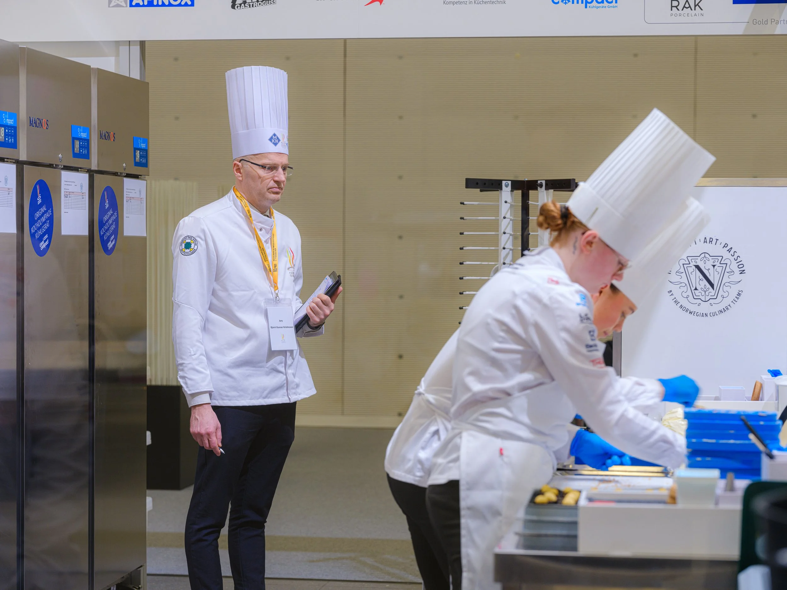 To kokker i hvite uniformer og kokkehatter jobber ved et arbeidsbord, mens en mann i bakgrunnen holder en telefon.