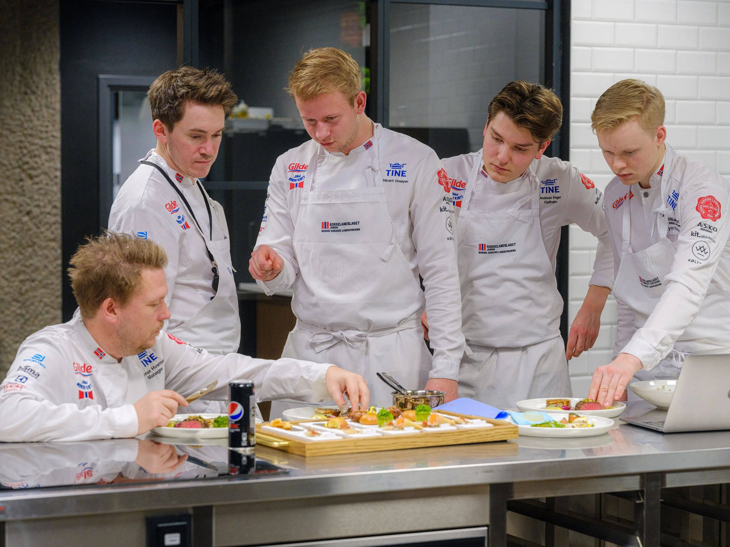 Fem unge kokkeklær undersøker og forbereder mat på et kjøkken.