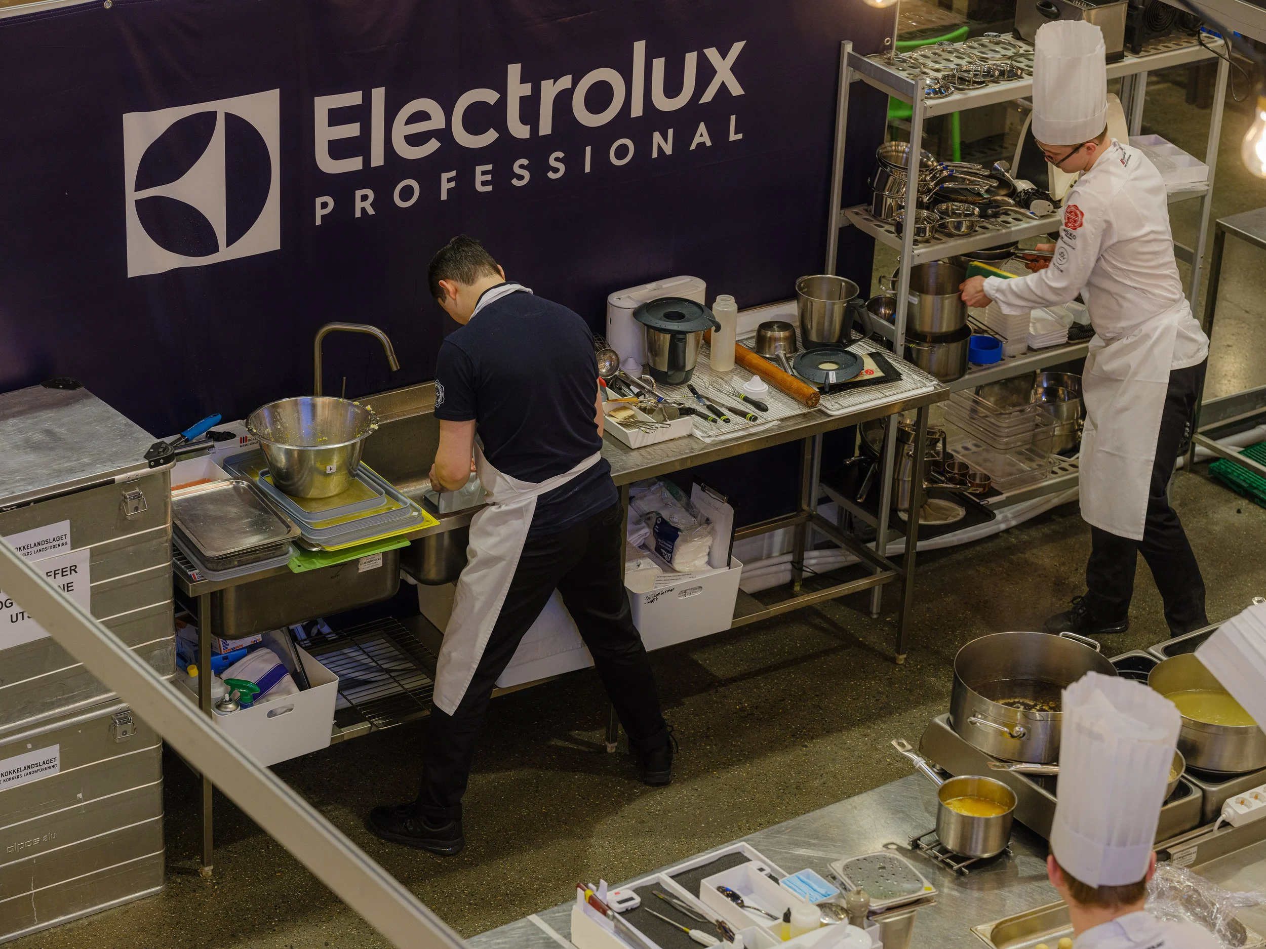 To kokker jobber på et profesjonelt kjøkken med Electrolux Professional-utstyr, en kok med hvit kokkehat, og et arbeidsbord med ulike kjøkkenredskaper og utstyr.