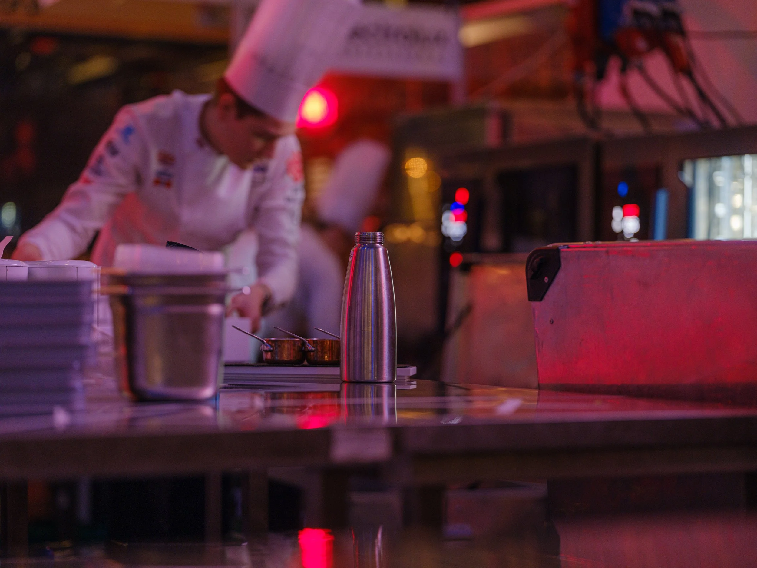 Kokk i hvit uniform og kokkehatt som jobber i et kjøkken, med fokus på arbeidsbenk og kjøkkenutstyr, med rosa og blå belysning.