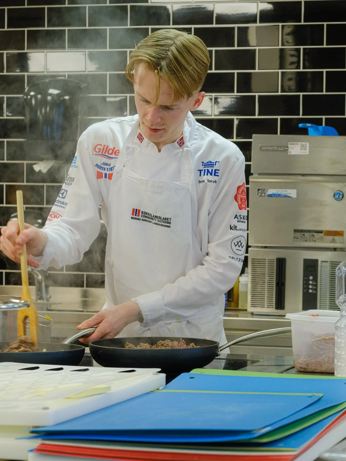 Kvinne med blondt, kuttet hår i kjøkken, steker kjøtt i stekepanne, omgitt av kjøkkenutstyr og fargerike benkeplater.