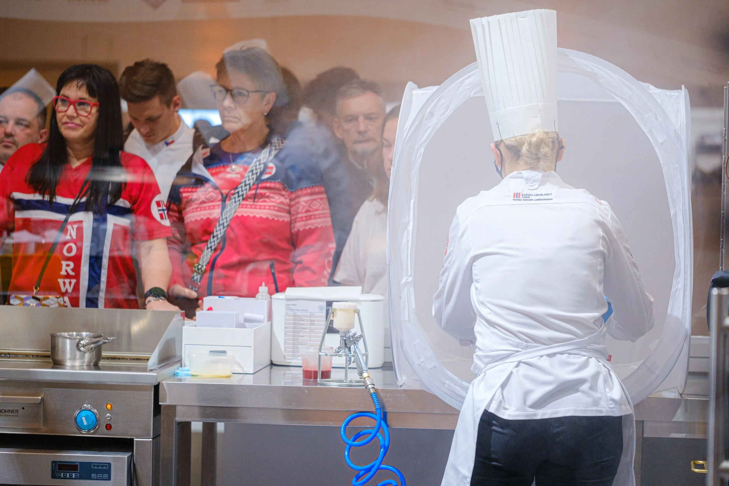 En kokk som bruker en medieskyddstelt, mens folk ser på den andre siden av en glassplate, med noen i bakgrunnen iført røde og blå klær.