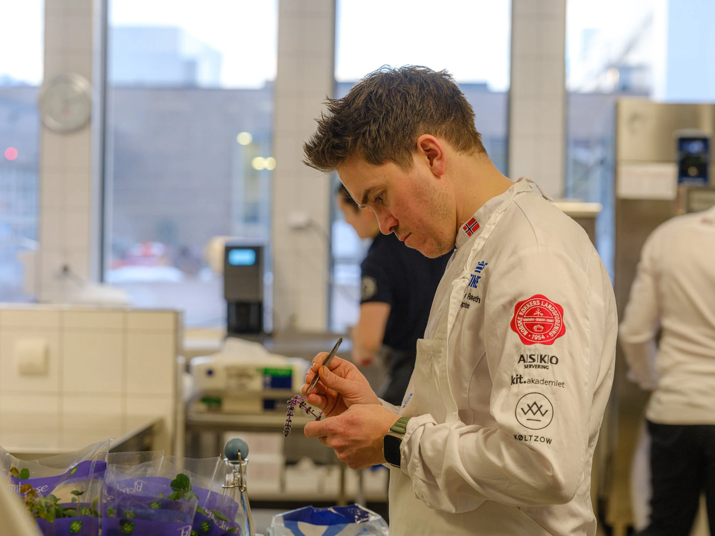 En mann i kokkelue skriver med en pinsett på en liten plante i et kjøkken.